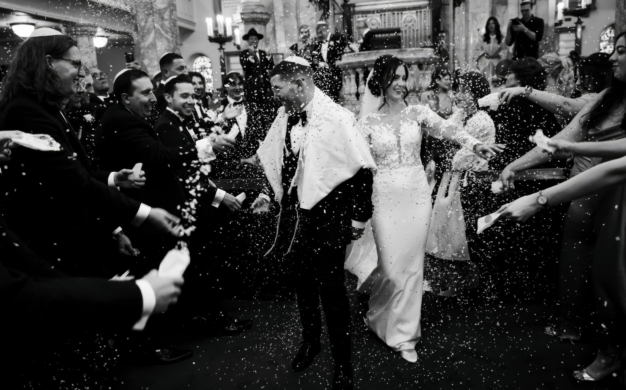 A black and white photo of a wedding celebration with a bride and groom walking through a confetti shower, surrounded by guests in a church or cathedral.