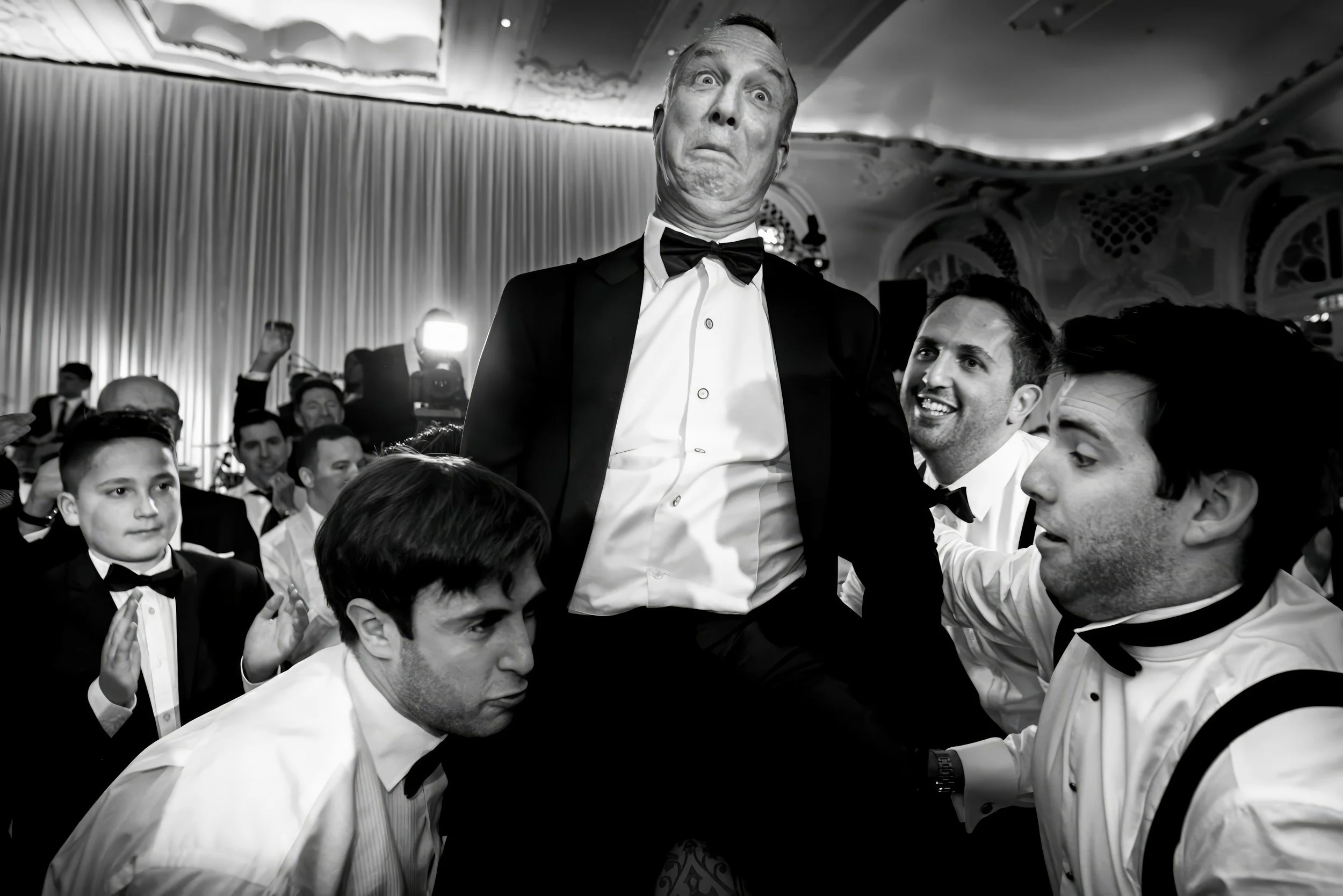 Black and white photo of a man in a tuxedo making a funny face, being lifted by four men in tuxedos at a formal event with a decorated ceiling and curtains in the background.