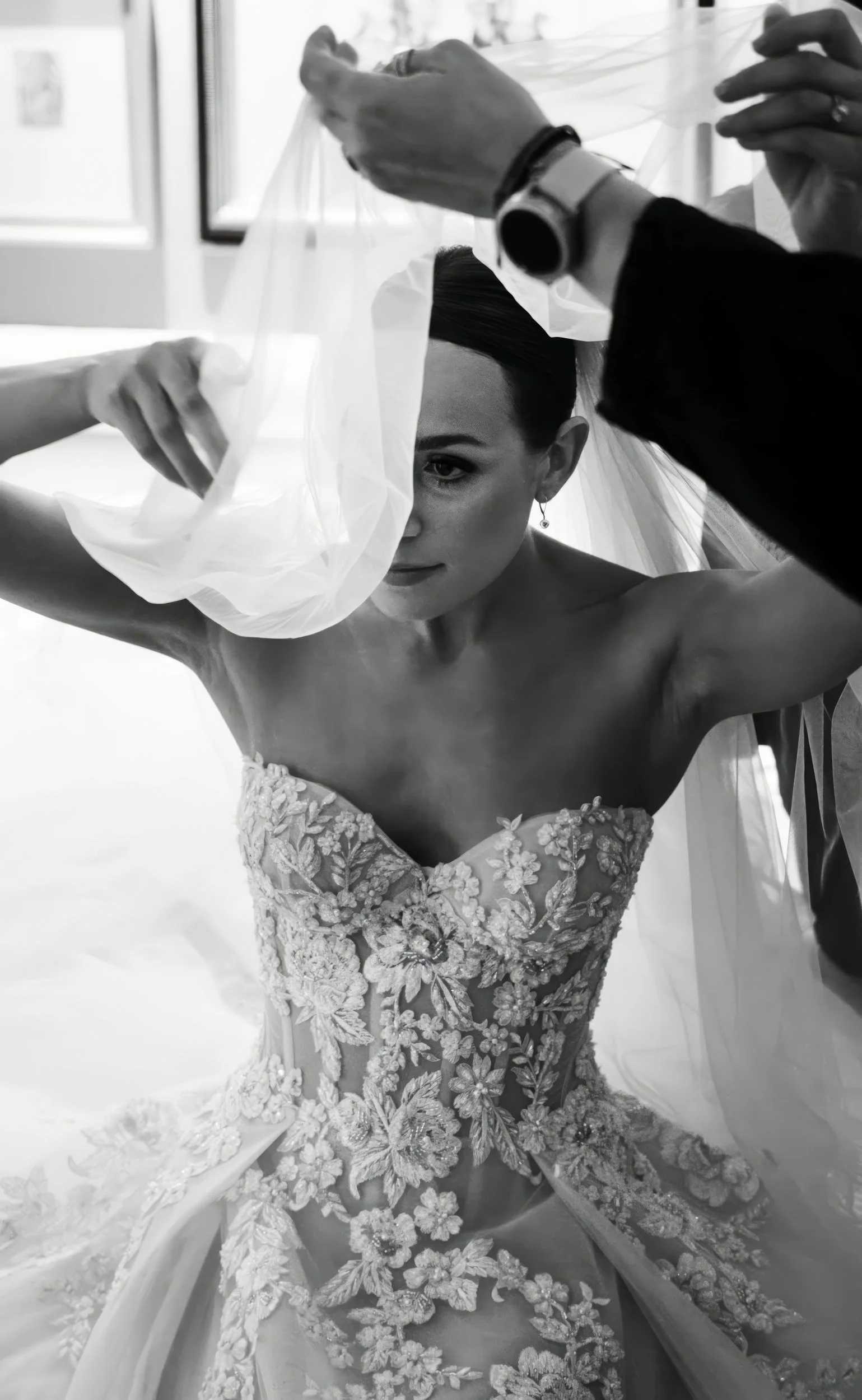 A bride in a strapless, embroidered wedding dress is getting her veil arranged with help from another person, likely a stylist or assistant, in a room with a window.