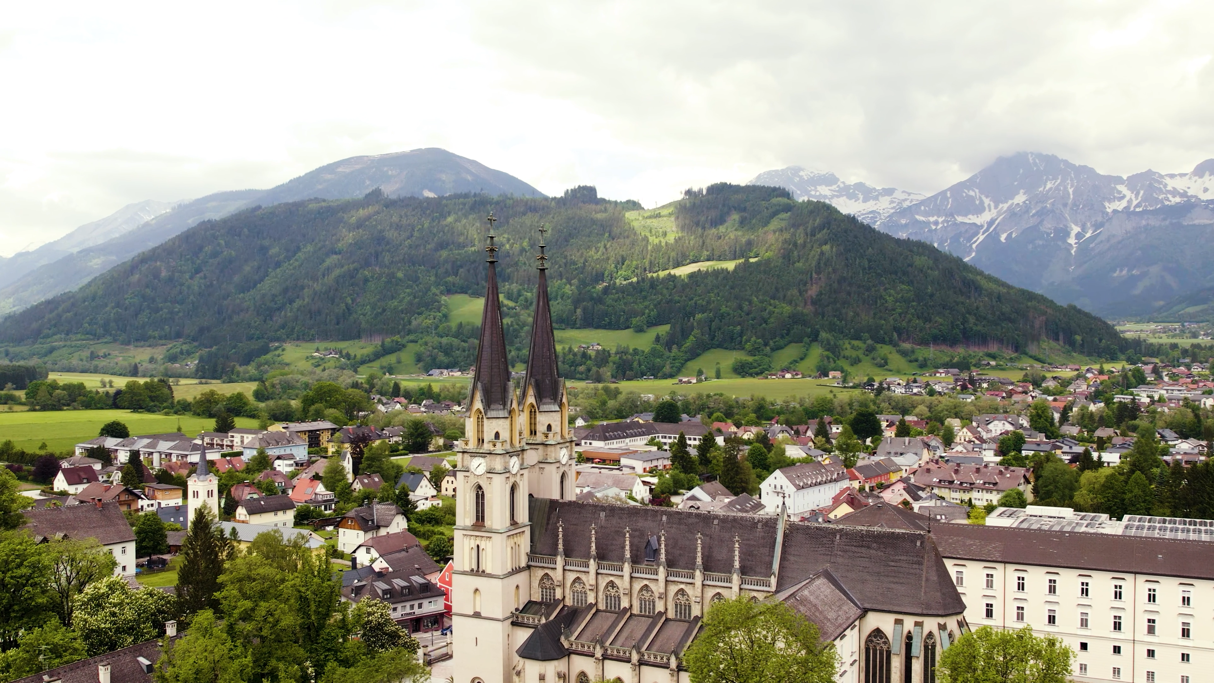 Aerial view of a town with a large church featuring two spires, surrounded by green hills and mountains in the background.