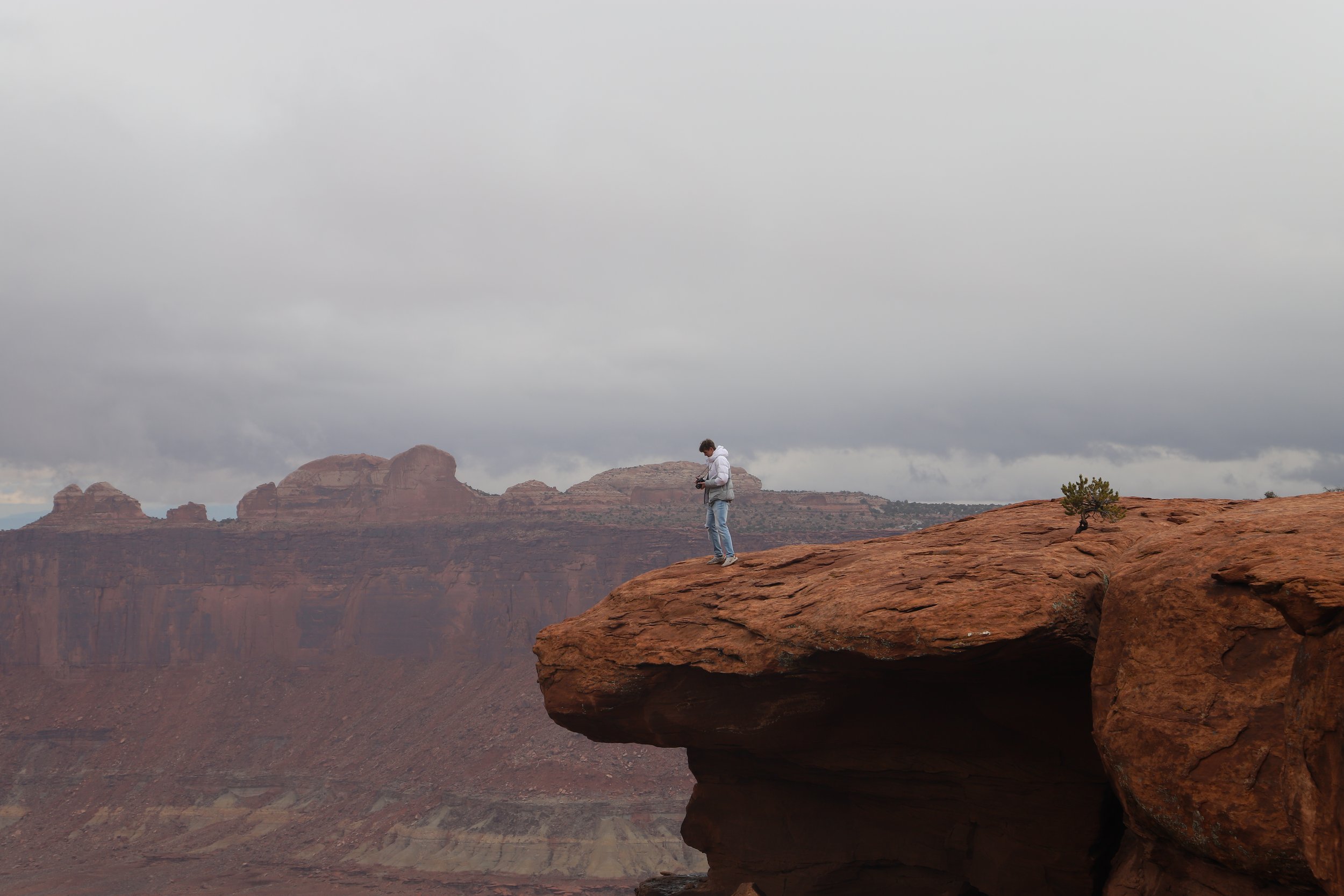Person standing on the edge of a rocky cliff with a camera, overlooking a vast canyon landscape under a cloudy sky.