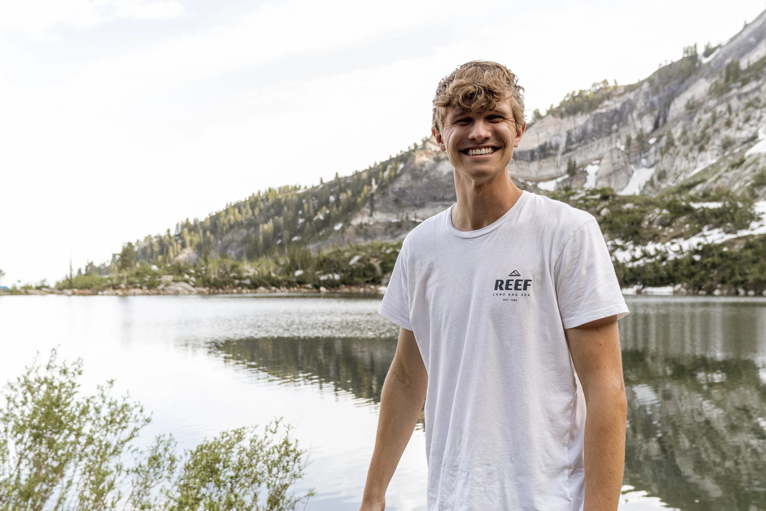Smiling person in a white t-shirt standing by a mountain lake with pine trees and rocky hills in the background.