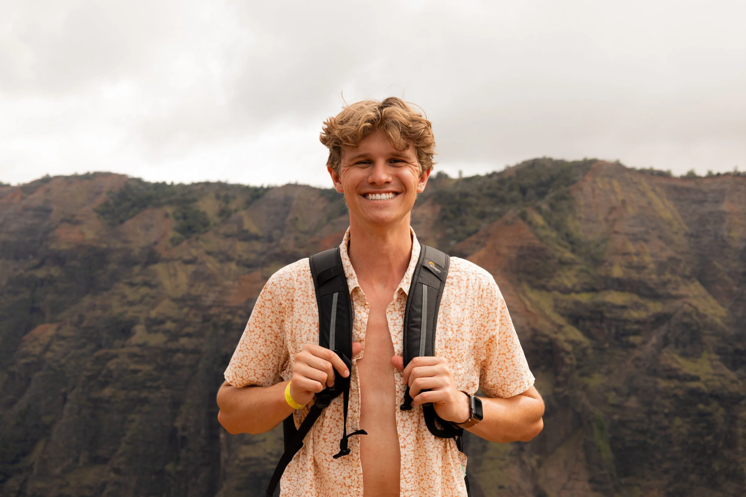 Person smiling outdoors with a backpack, mountain scenery in the background.