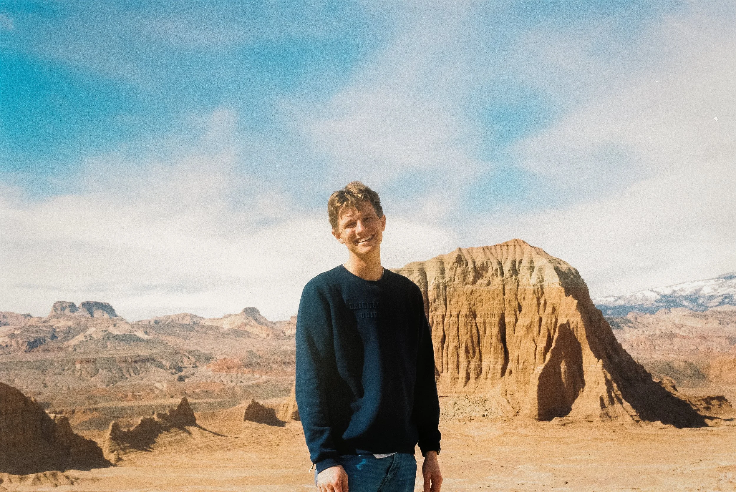 Person smiling in front of a desert landscape with rock formations and a blue sky.