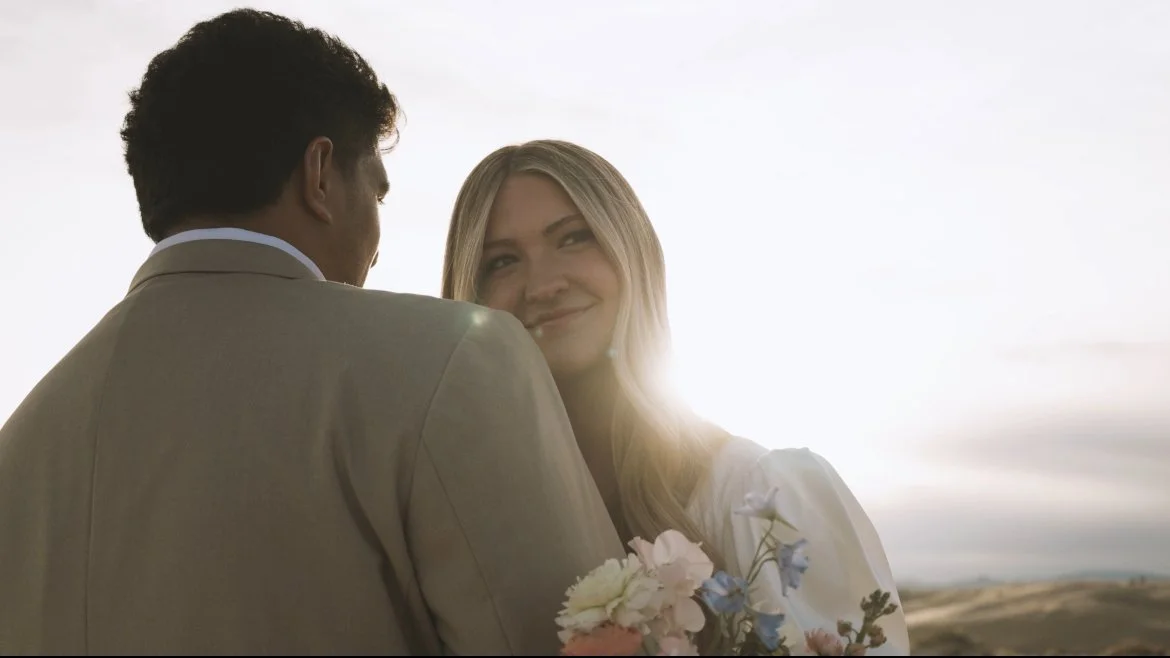 A man and woman embracing outdoors with the sun setting in the background, the woman smiling and holding a bouquet.