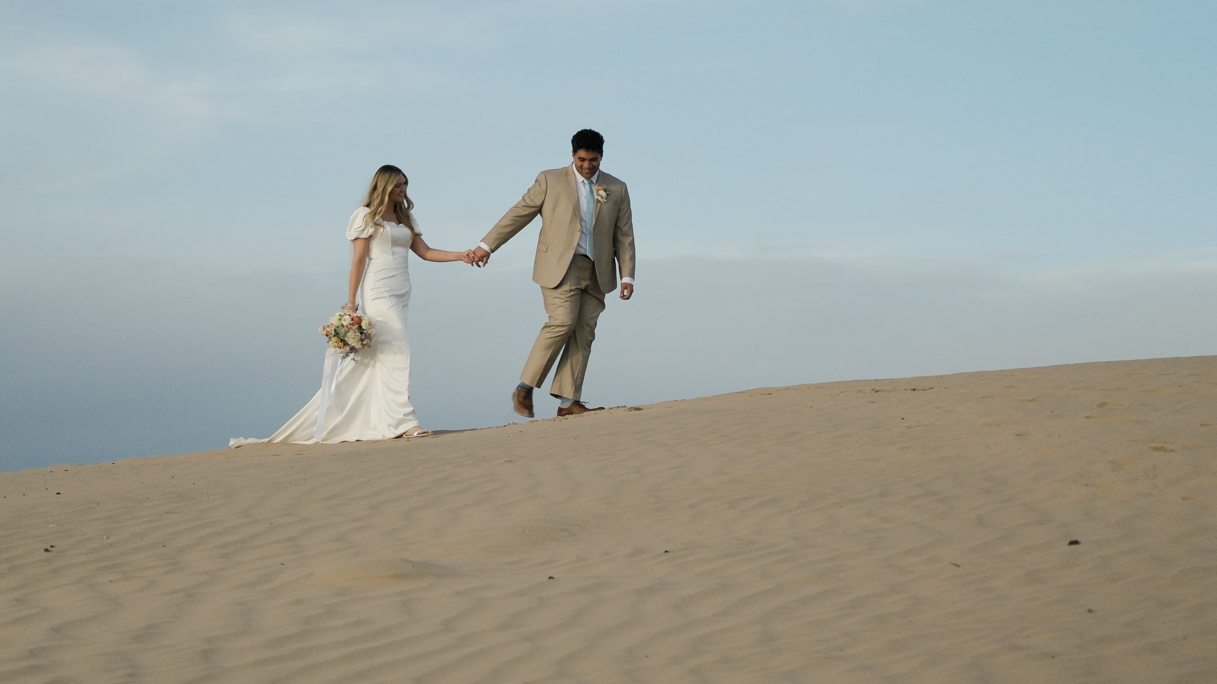 Couple walking on a sandy hill in wedding attire holding hands.