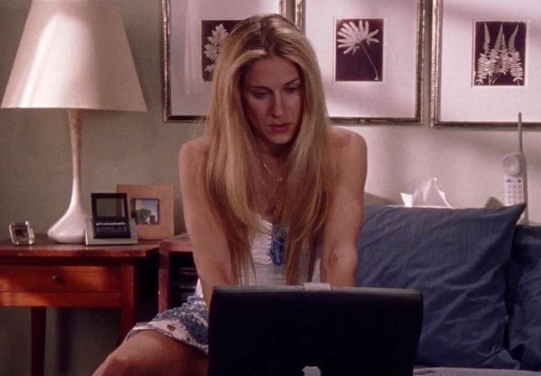 A woman with long blonde hair sitting on a bed, looking at a laptop. Behind her are framed botanical prints and a nightstand with a lamp, a picture frame, and other items.