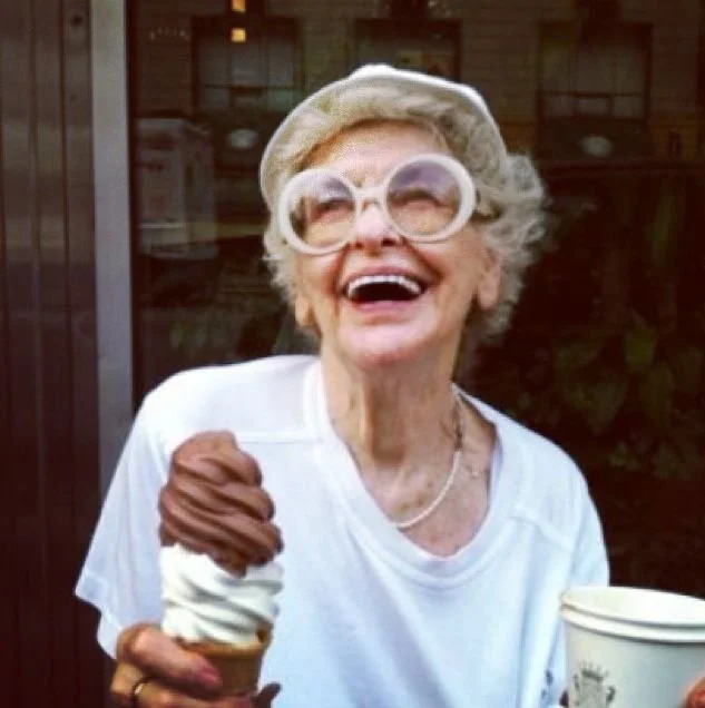 An elderly woman with white hair, large round glasses, and a white hat laughing outdoors while holding an ice cream cone with vanilla and chocolate ice cream. She is happy regardless of what others think.