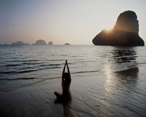 A person practicing yoga on the beach at sunset with a large rock formation in the background.