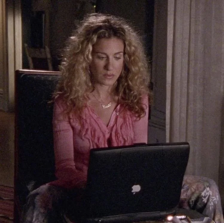 A woman with curly blonde hair sitting in a dark room, using a black laptop, wearing a pink top.
