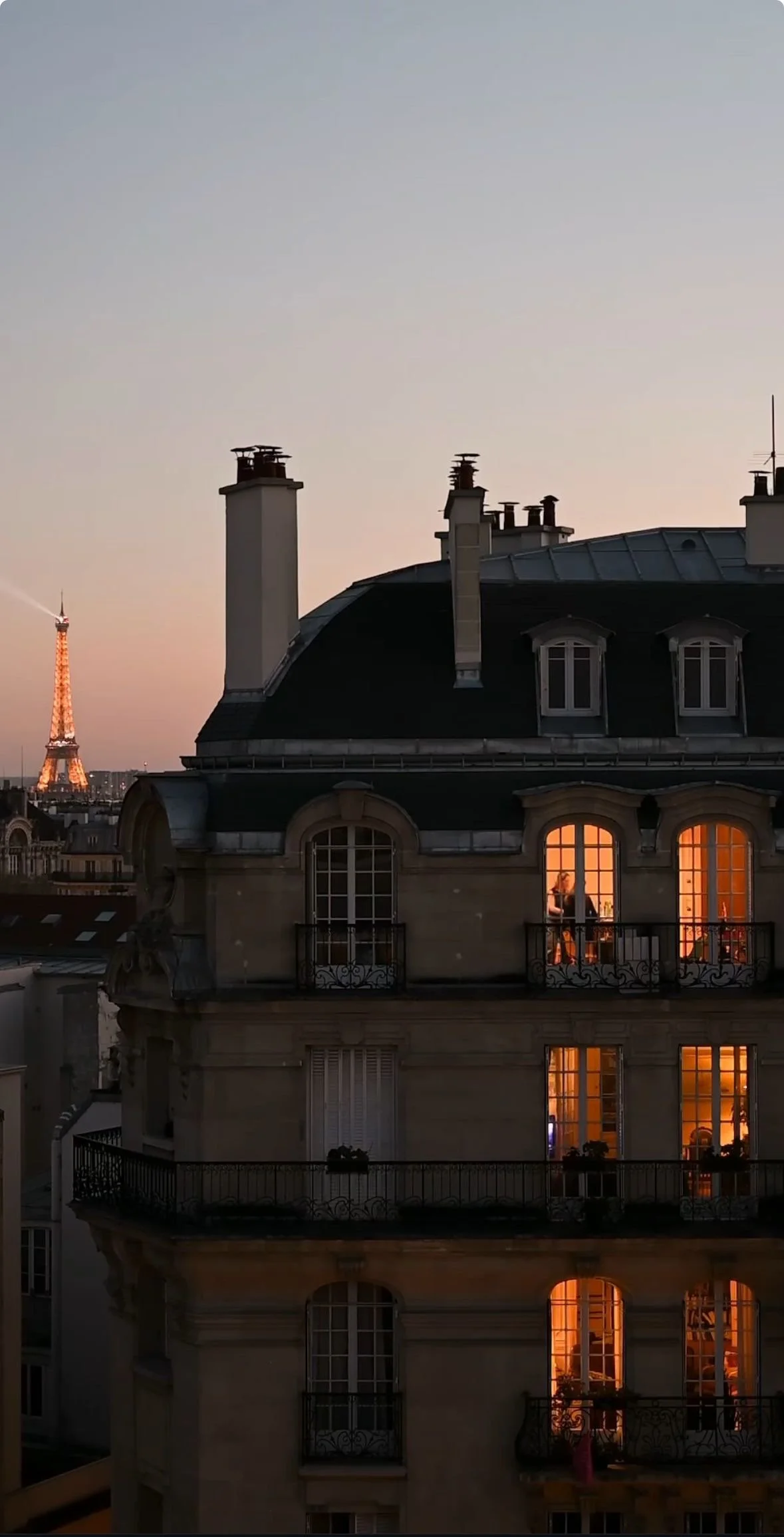 Parisian apartment building at sunset, with interior lights visible through large windows and the Eiffel Tower illuminated in the background.