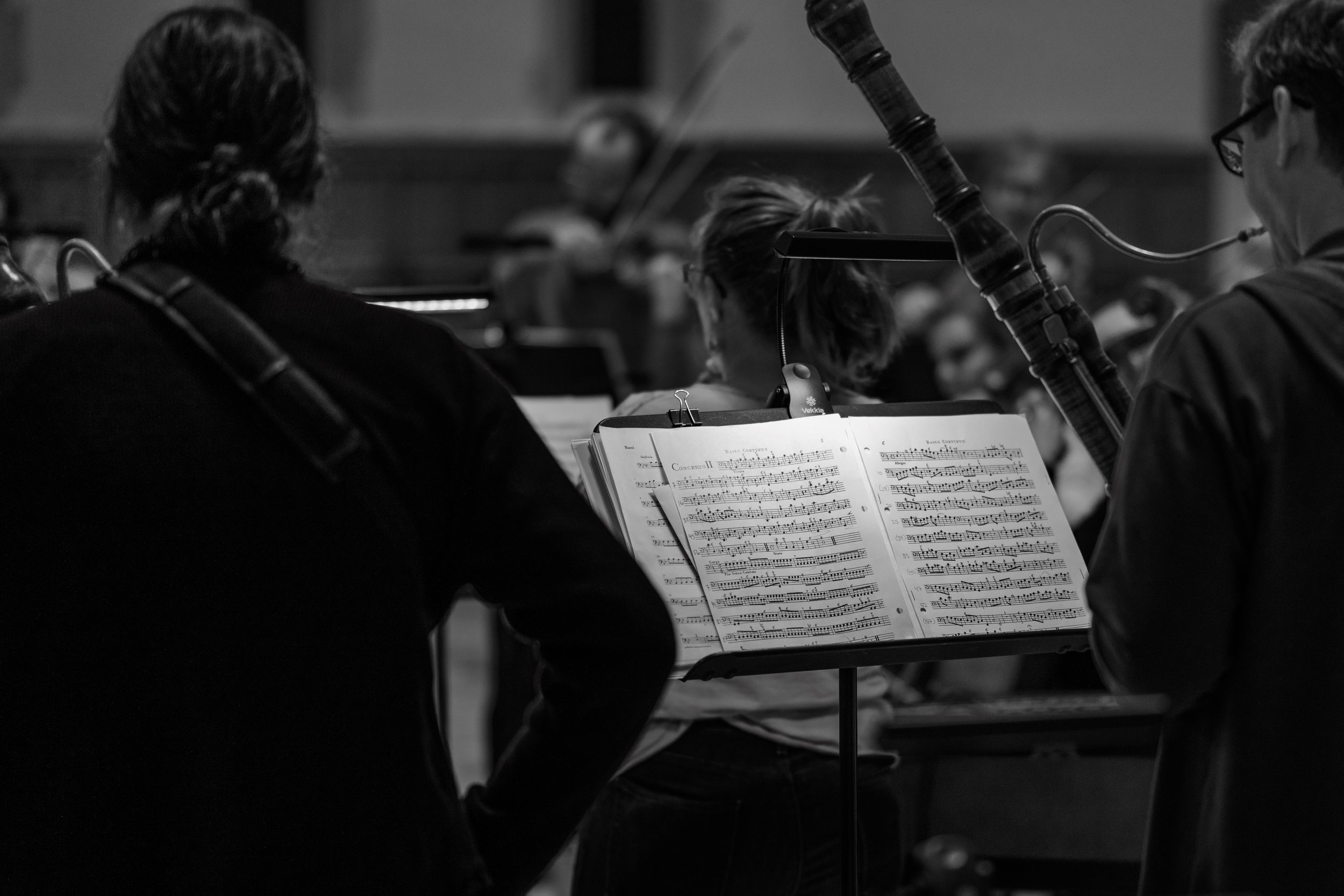 Musicians in an orchestra rehearsing, with sheet music on stands, in a black-and-white photo.