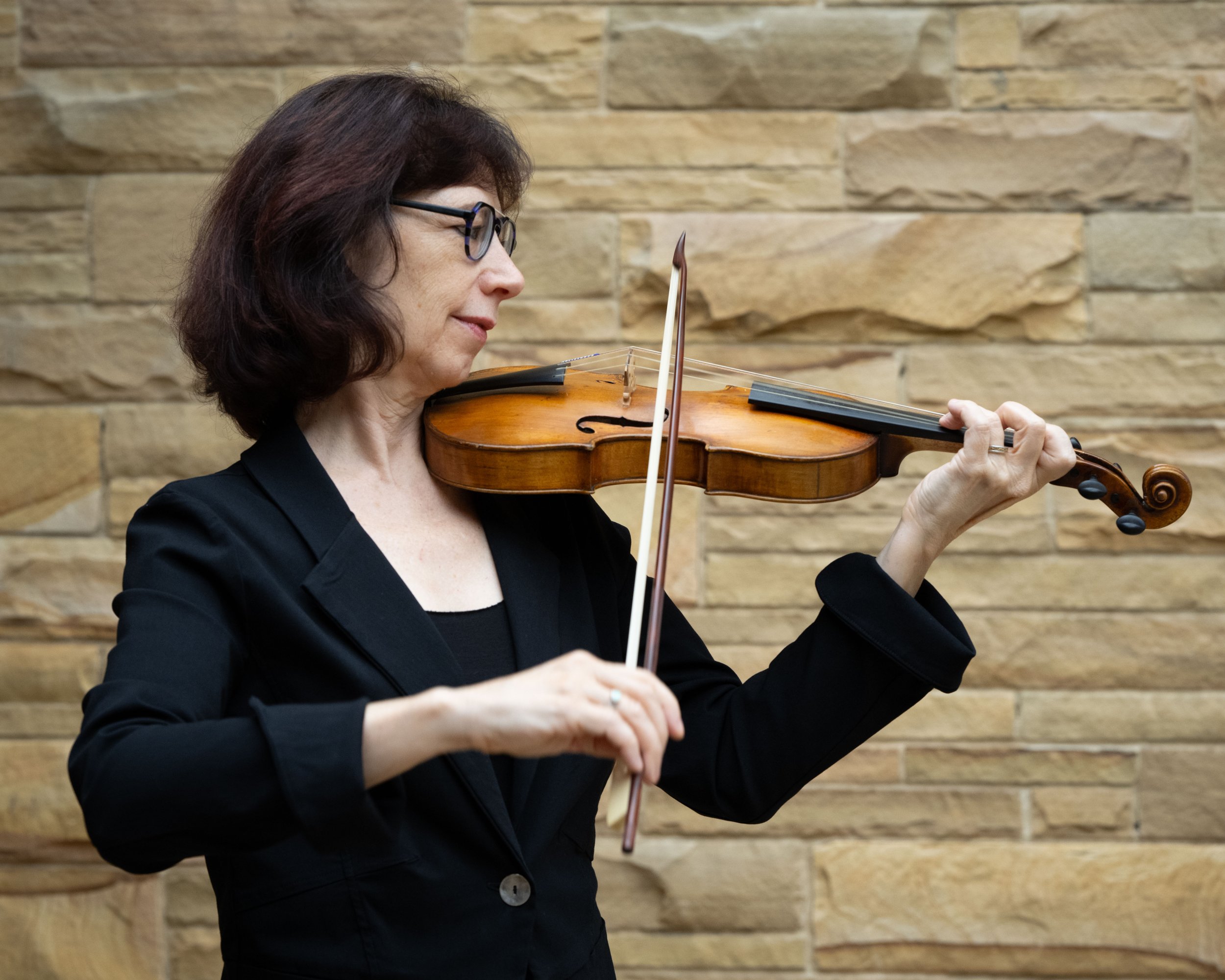 A woman with glasses playing a violin against a brick wall background.
