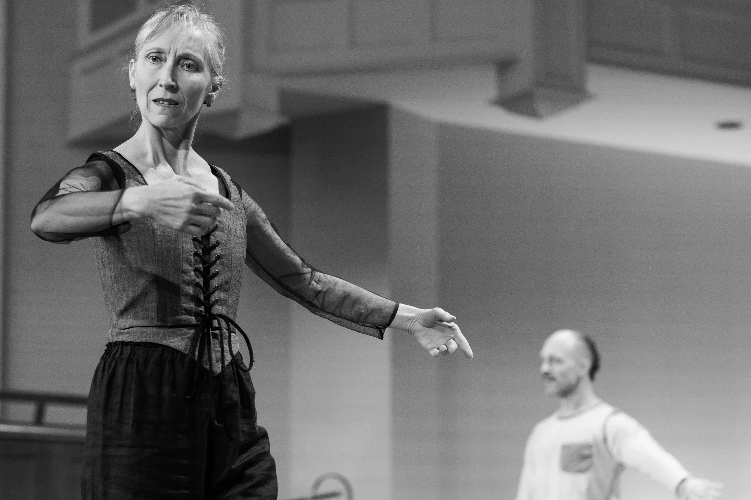 A woman with gray hair in dance attire, pointing with her right hand, appears to be instructing or demonstrating. A man with a beard and glasses in casual attire stands behind her, smiling with his arms spread out.