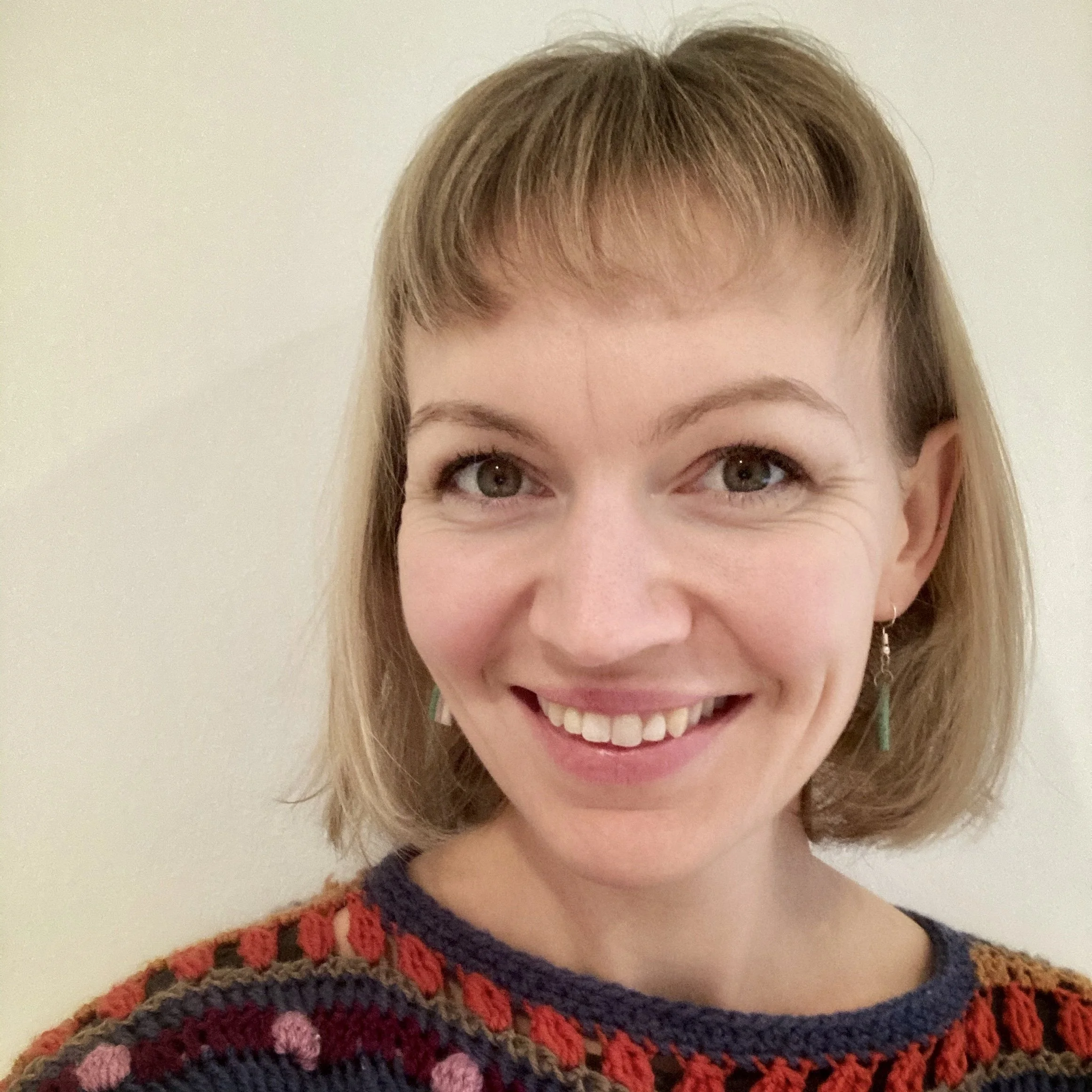 A smiling woman with light brown hair and green eyes, wearing earrings and a colorful knit sweater, standing against a plain light-colored wall.