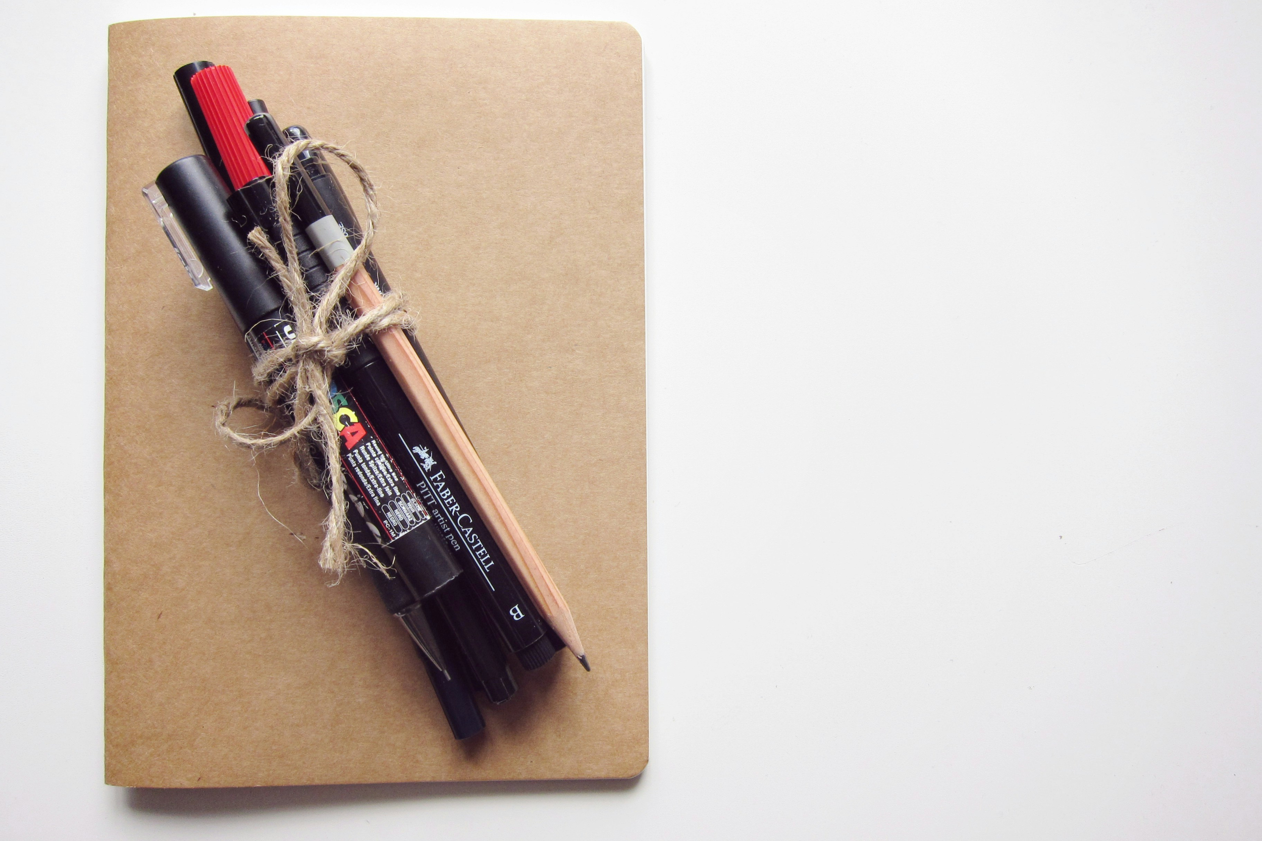 A brown notebook with a bundle of black, red, and brown drawing pens and a wooden pencil tied together with a piece of twine, placed on a white surface.