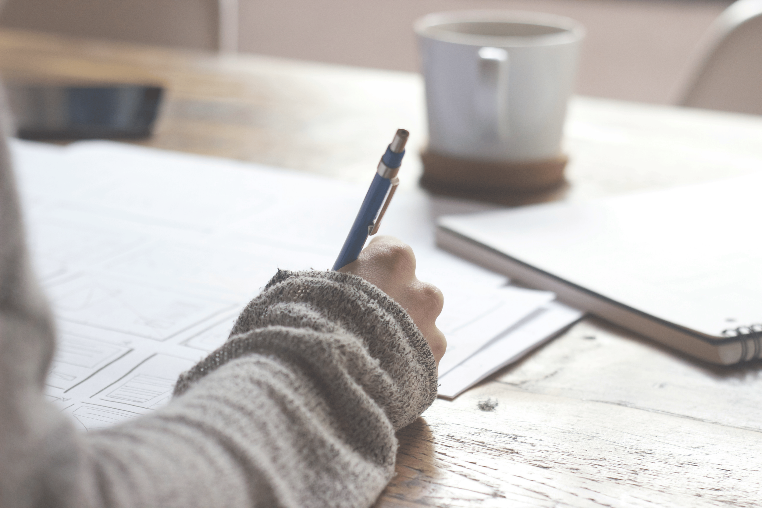 A person wearing a gray, fuzzy sweater writing on papers at a wooden table, with a notebook and a mug in the background.