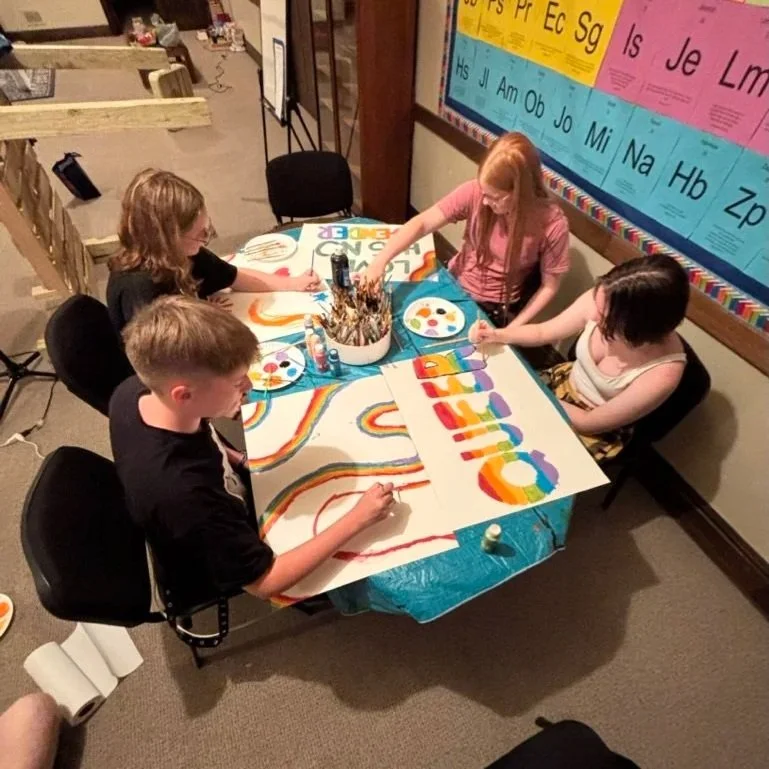 Four children seated around a table painting a colorful rainbow-themed poster.