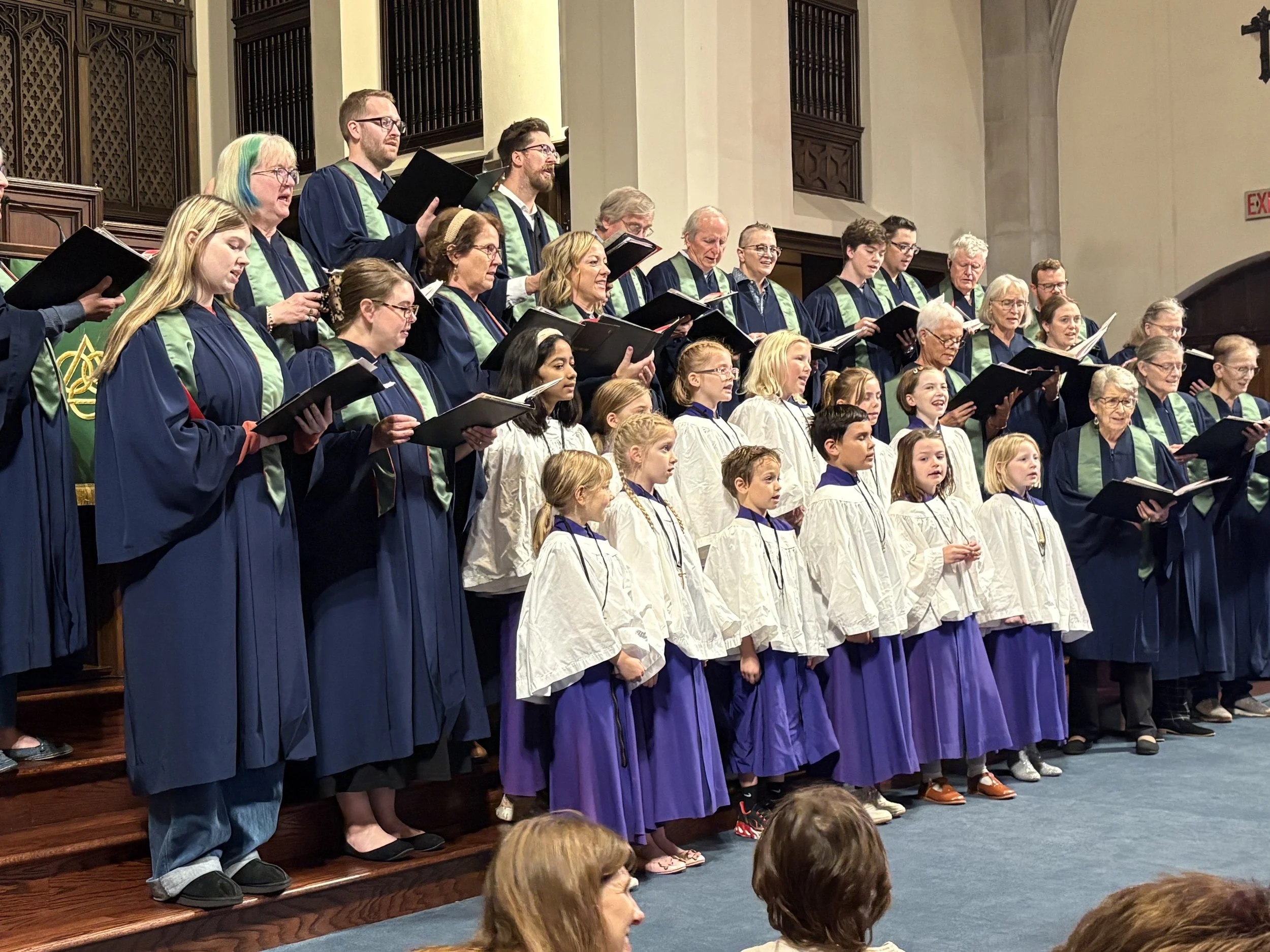 A choir of adults and children singing together on stage in a church or concert hall, dressed in robes and holding sheet music.
