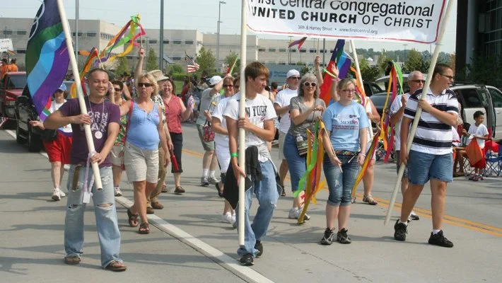 A procession of people marching outdoors holding rainbow flags and banners, including a sign for First Central Congregational United Church of Christ