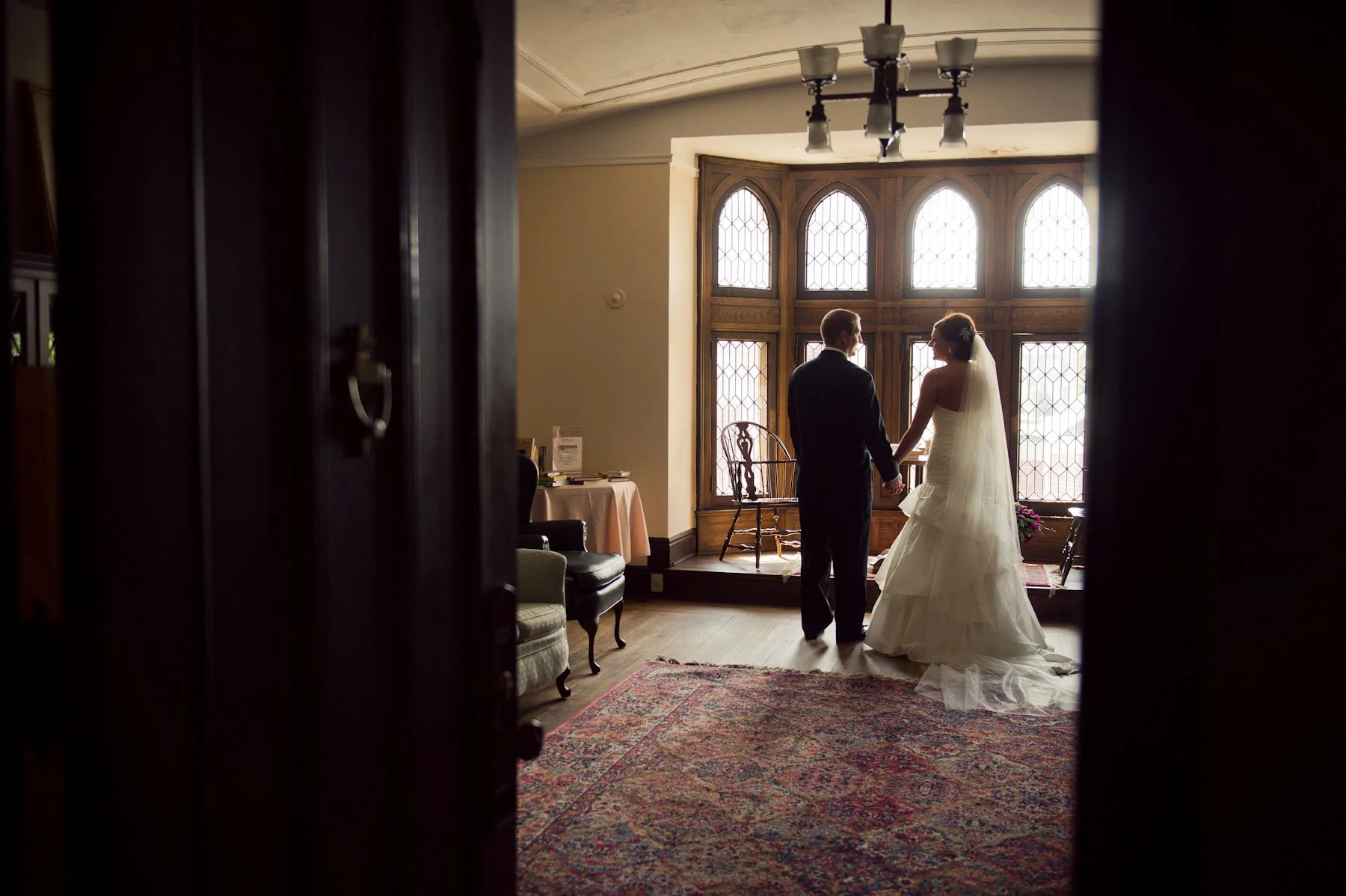 A bride and groom holding hands in a room with large stained glass windows, a chandelier, and vintage furniture, seen through an open door.