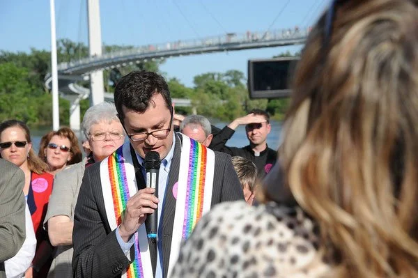 A man in a suit with a rainbow pride sash speaking into a microphone at an outdoor event with a crowd of people, some wearing sunglasses, and a bridge in the background.