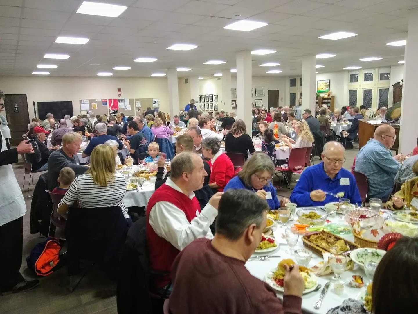 A large indoor gathering of people seated at round banquet tables, enjoying a meal, in a community hall with white walls, ceiling lights, and framed pictures.