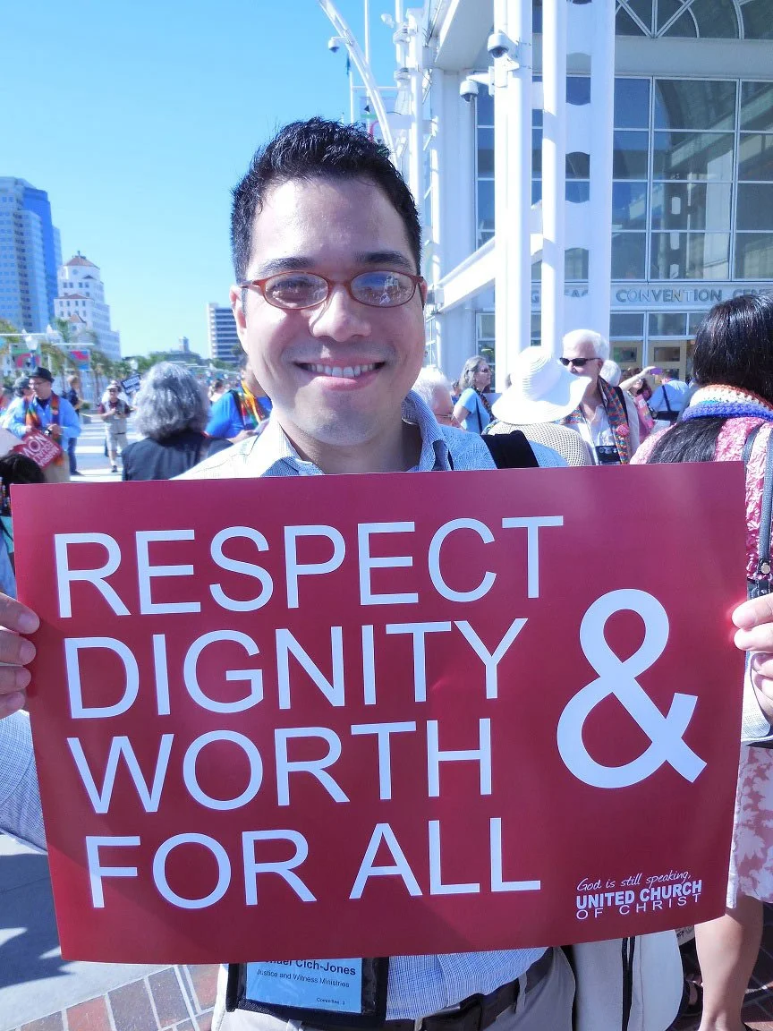 A smiling man holding a pink sign that reads 'Respect Dignity & Worth For All' at an outdoor gathering or rally, with other people and a cityscape in the background.