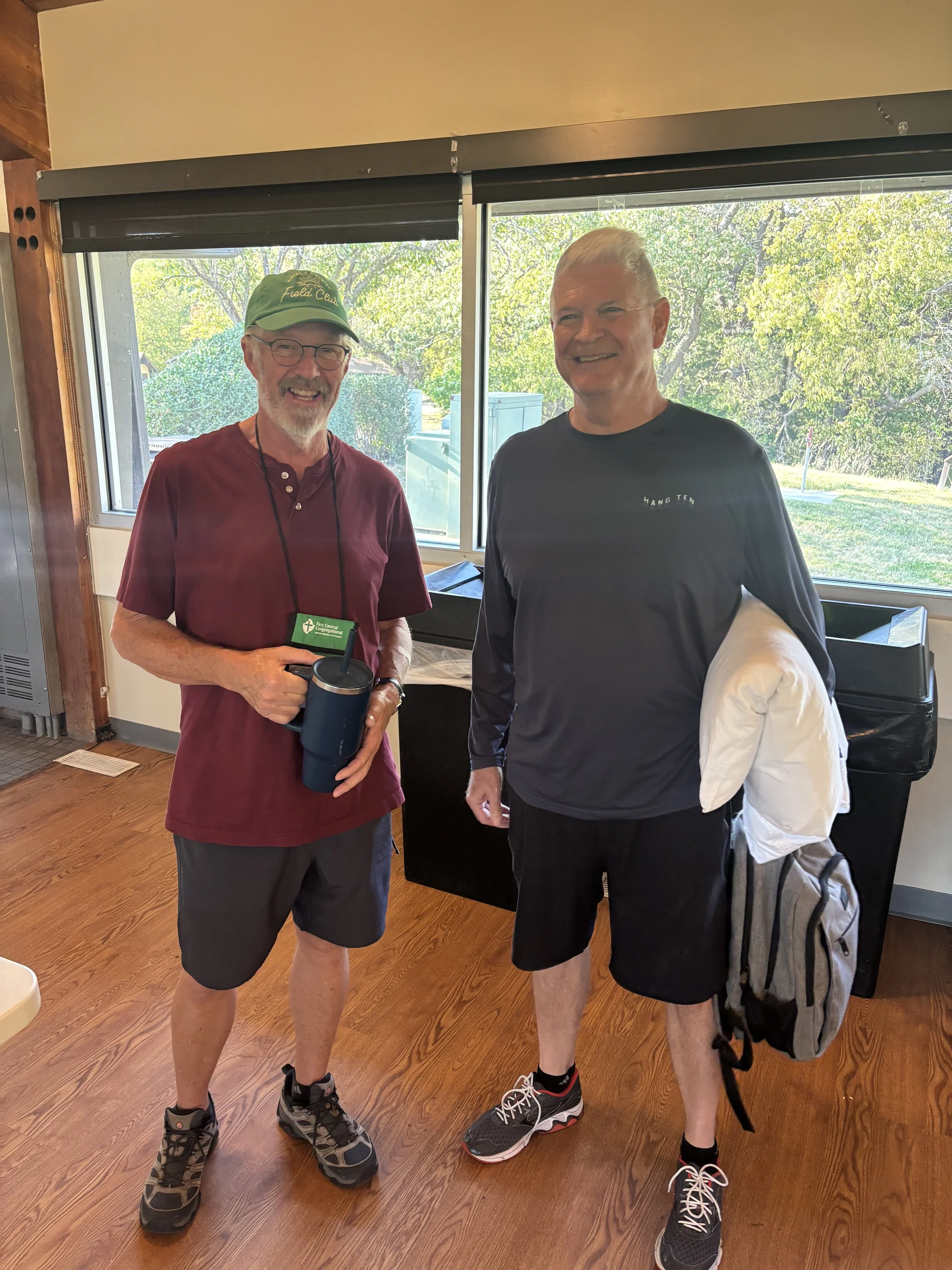Two smiling middle-aged men standing inside near a large window with green trees outside. One is wearing a maroon shirt, shorts, sneakers, and a baseball cap, holding a tumbler. The other is in a black long-sleeve shirt, shorts, black sneakers, and holding a backpack and a white jacket.