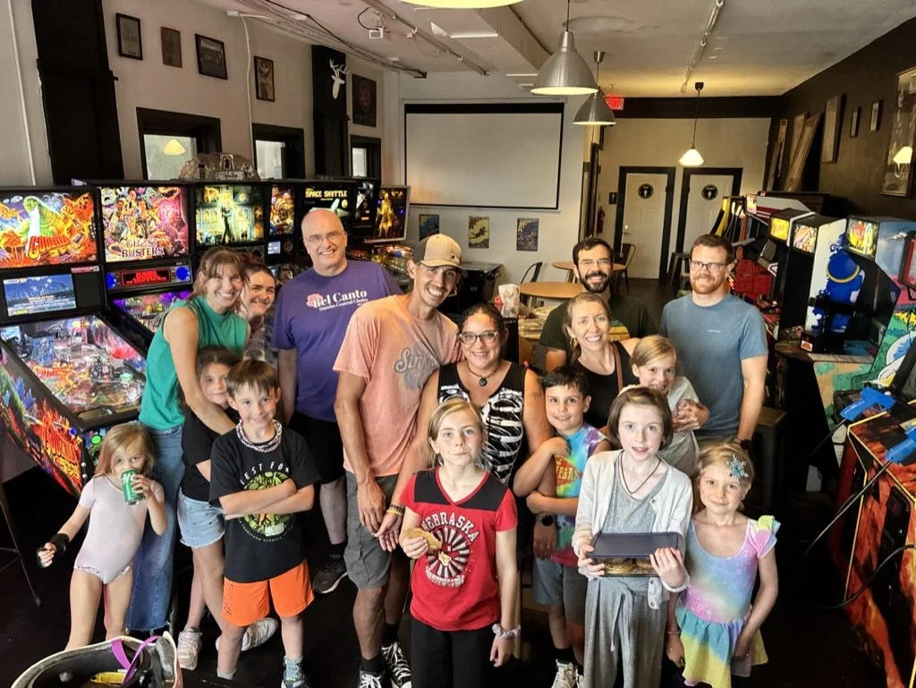 Group of children and adults in an arcade, posing for a photo with pinball machines and arcade games in the background.