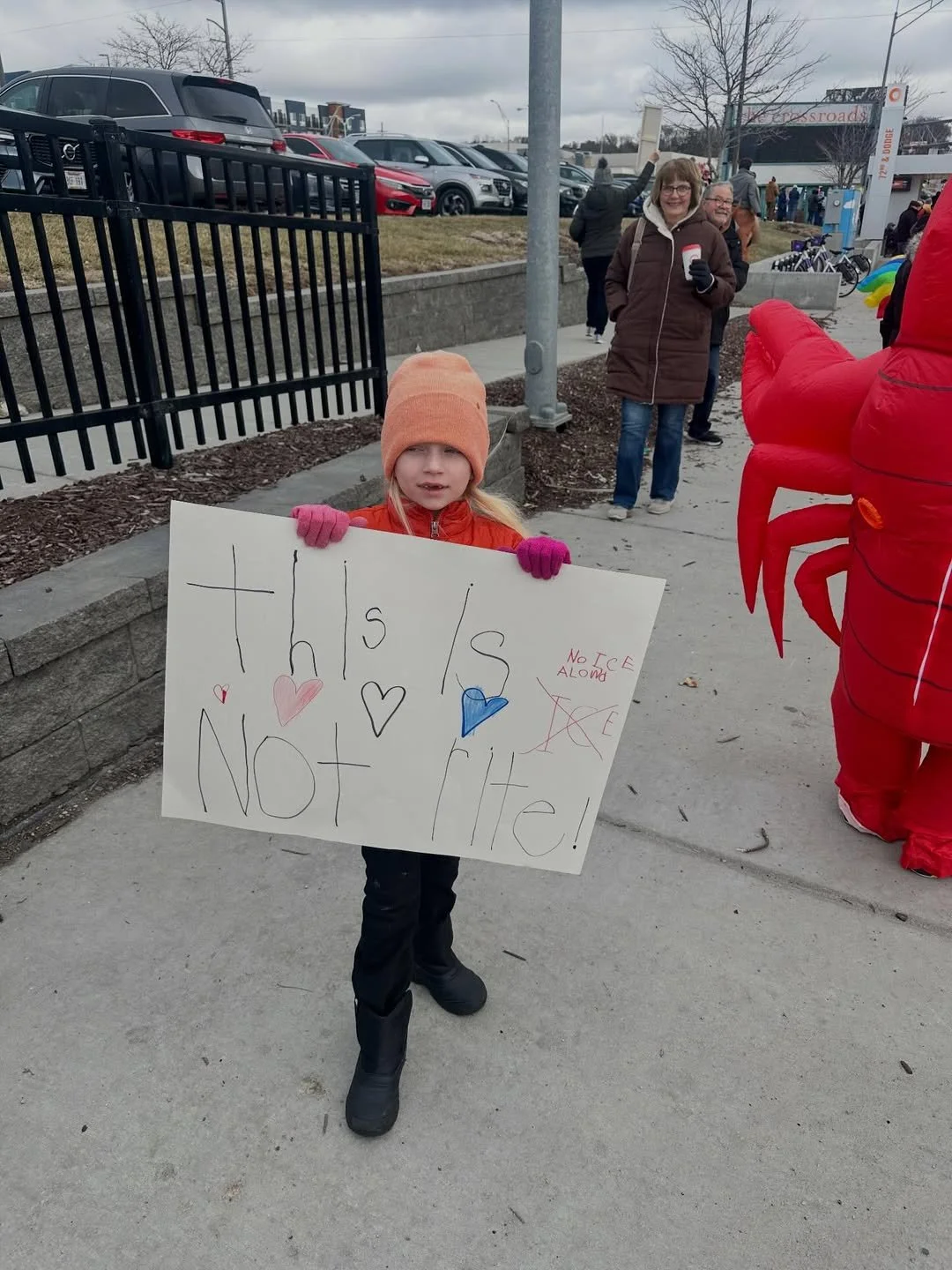 A young girl wearing a peach-colored beanie, pink gloves, and a red jacket holding a sign that says 'This is not right!' with hearts and a note in red ink saying 'No ice along' during a protest or rally. People and cars are visible in the background, and some individuals are holding signs or balloons.