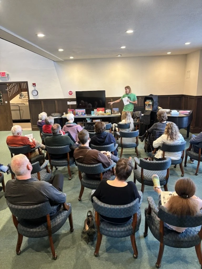 A woman giving a presentation to a group of elderly people seated in a room. The woman is holding papers and gesturing towards a large TV.