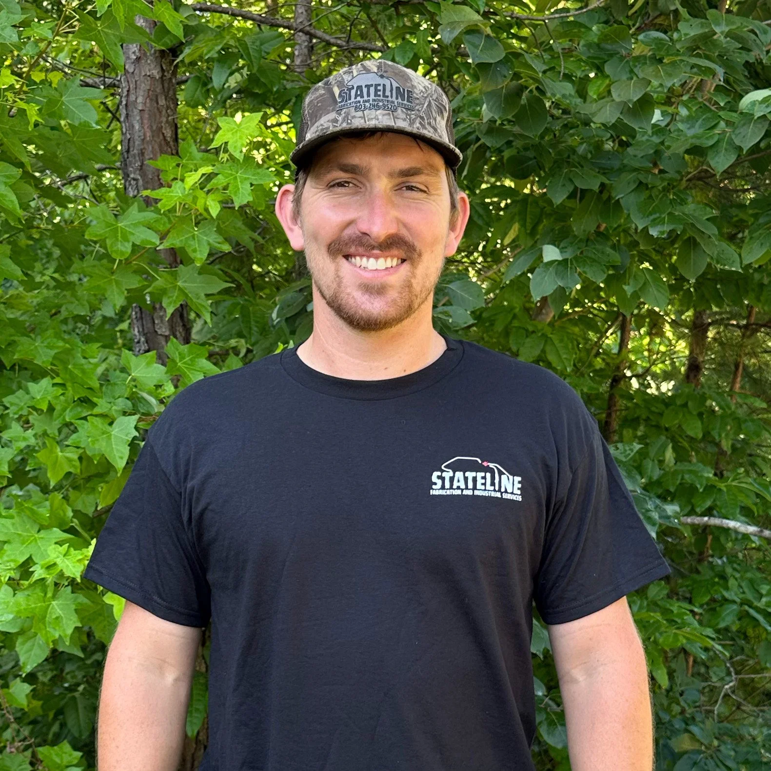 Augie Peterson, wearing a black Stateline Fabrication tshirt, smiling in front of a background of green trees
