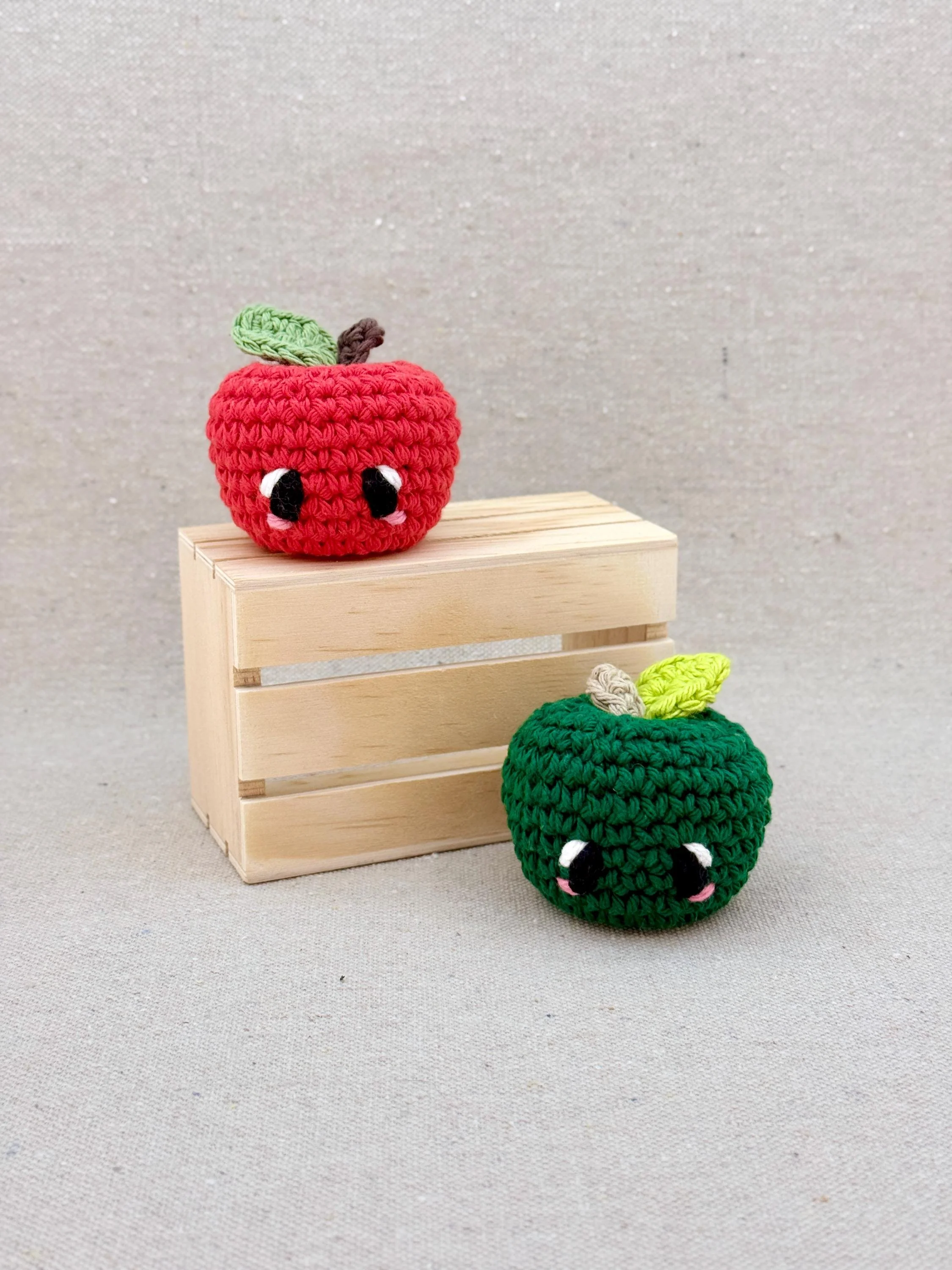 Two crochet amigurumi apple plushies posed with a mini wooden crate in red and pine green