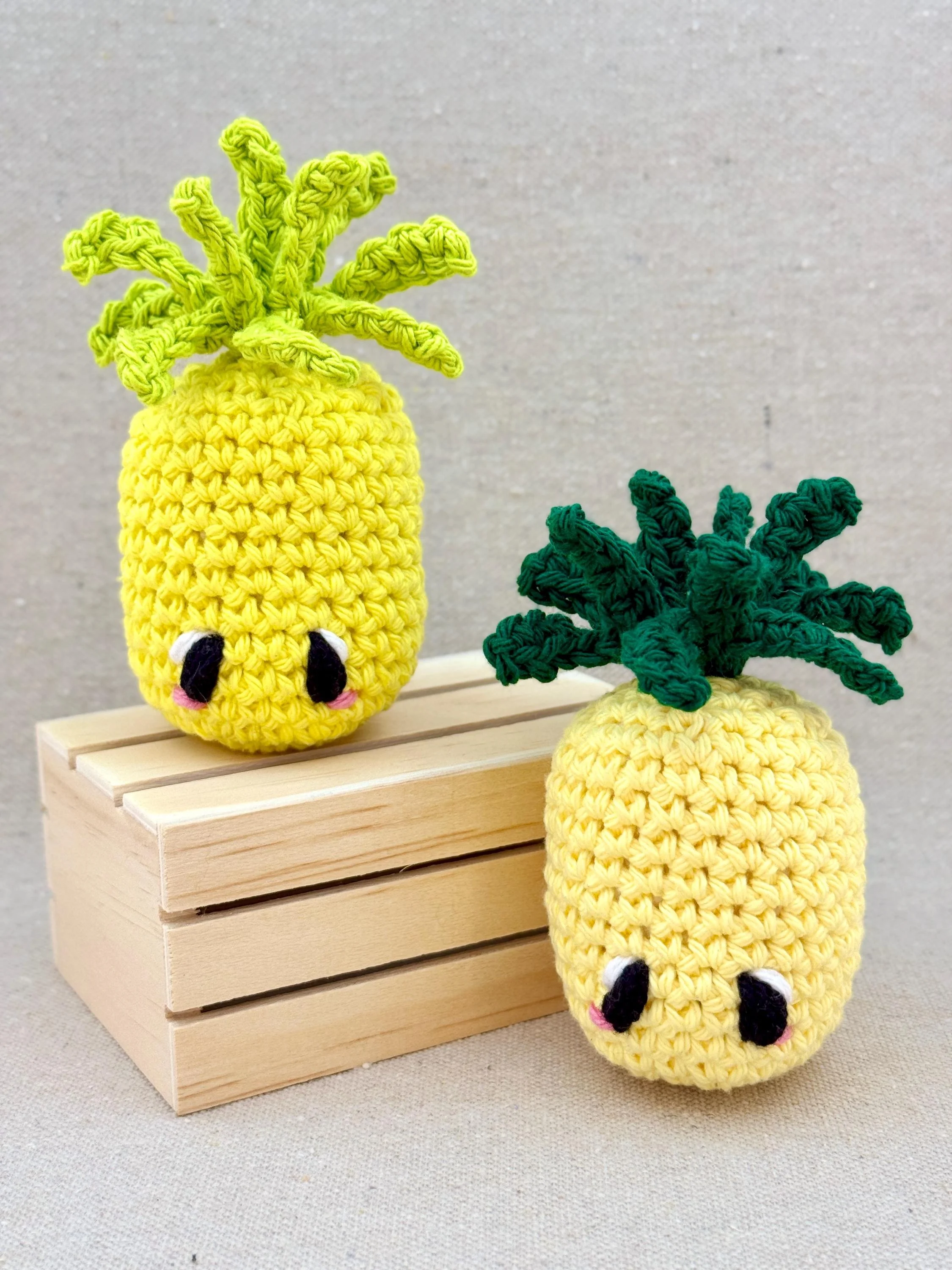 Two crochet amigurumi pineapple plushies posed next to a mini wooden crate in bright yellow and butter yellow (Copy)