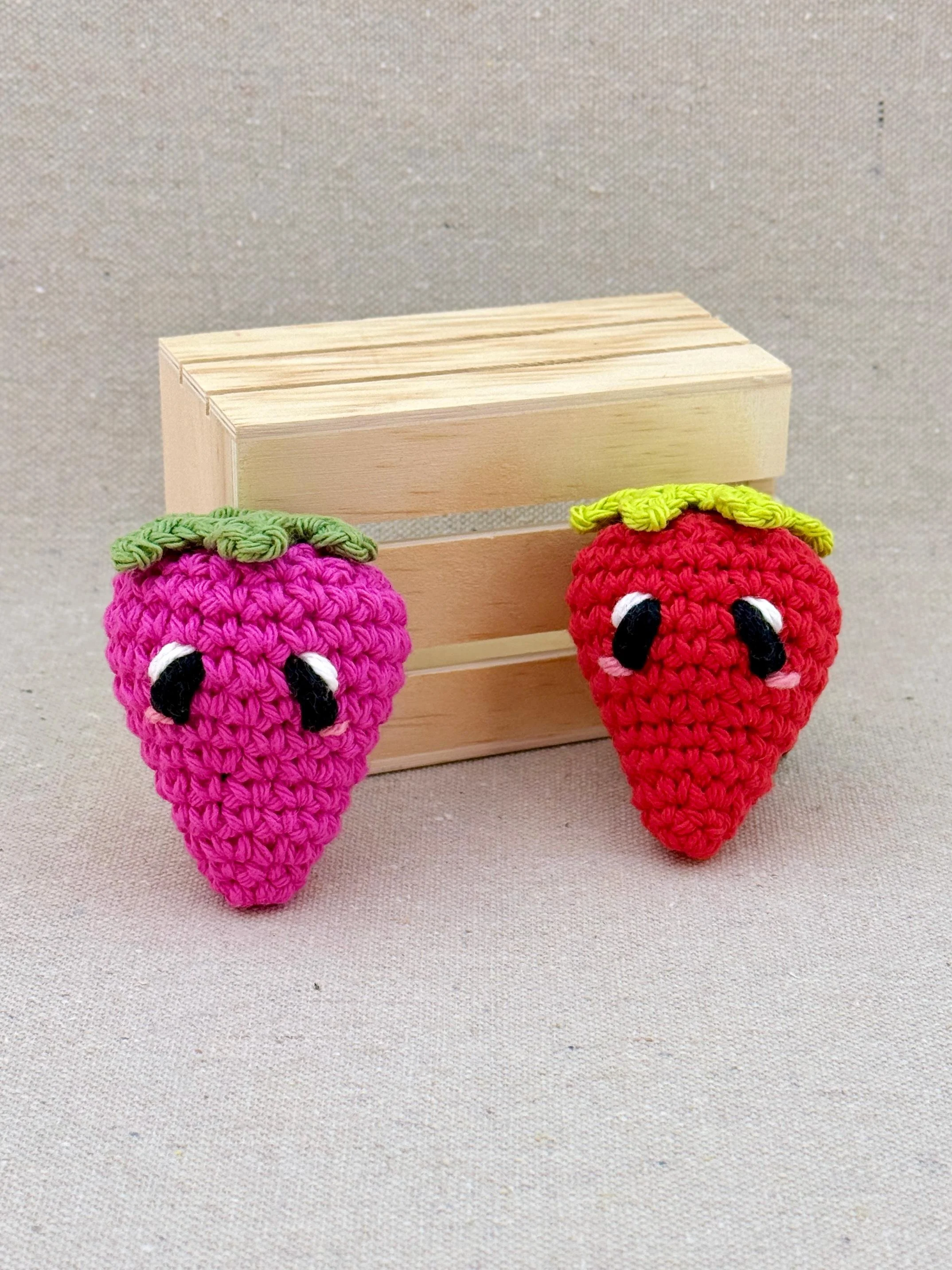 Two crochet amigurumi strawberry plushies posed next to a mini wooden crate in red and hot pink (Copy)