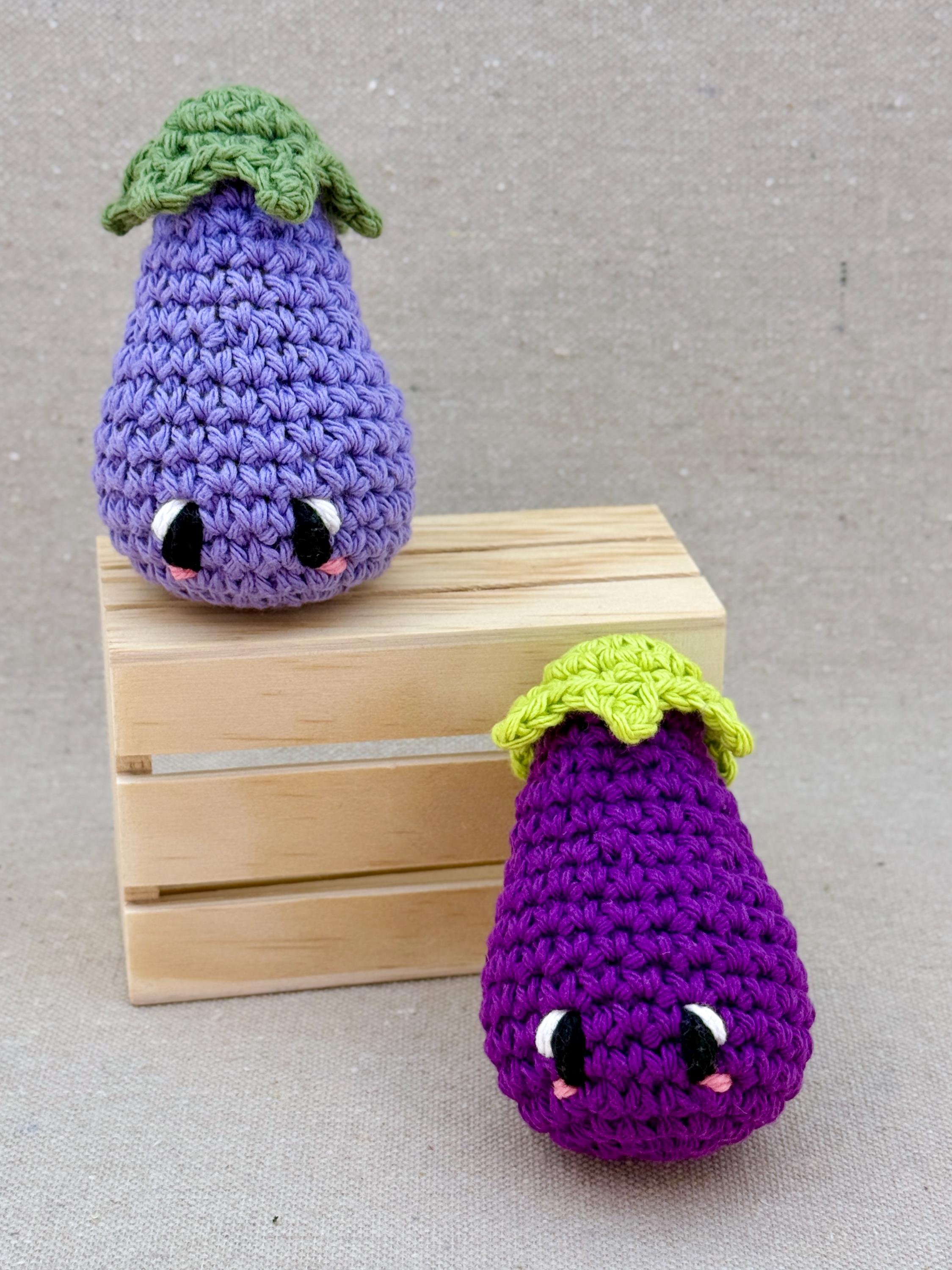 Two crochet amigurumi eggplant plushies posed with a mini wooden crate in dark purple and light purple