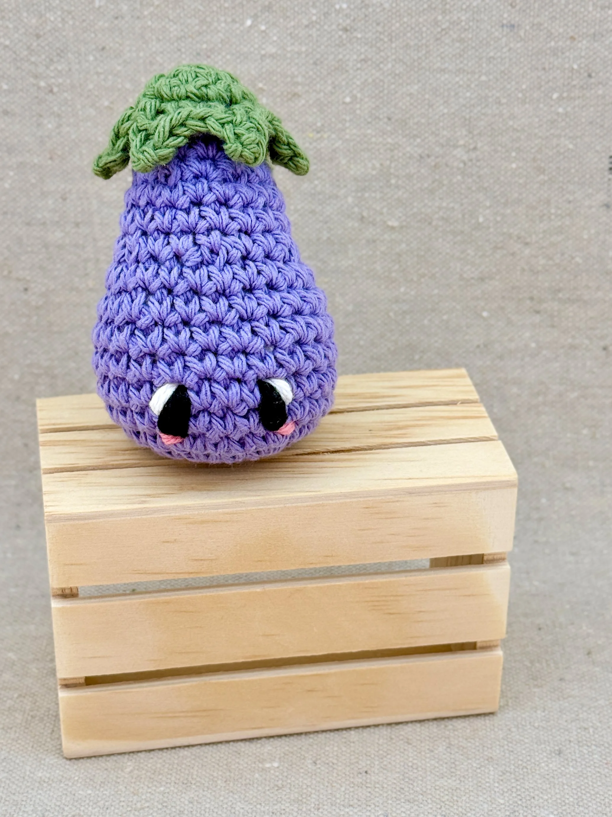 A light purple amigurumi eggplant plush posed with a mini wooden crate