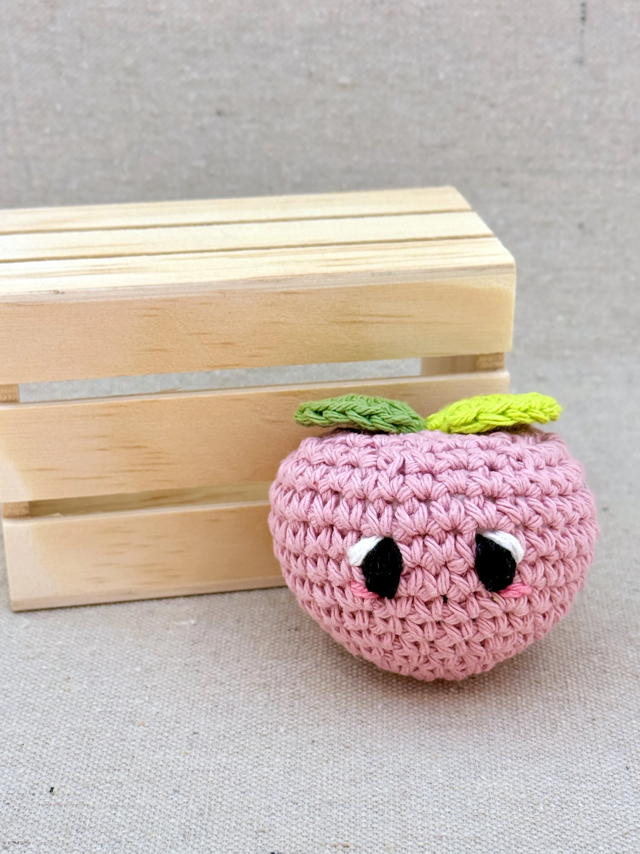 A bubblegum amigurumi peach plush posed next to a mini wooden crate