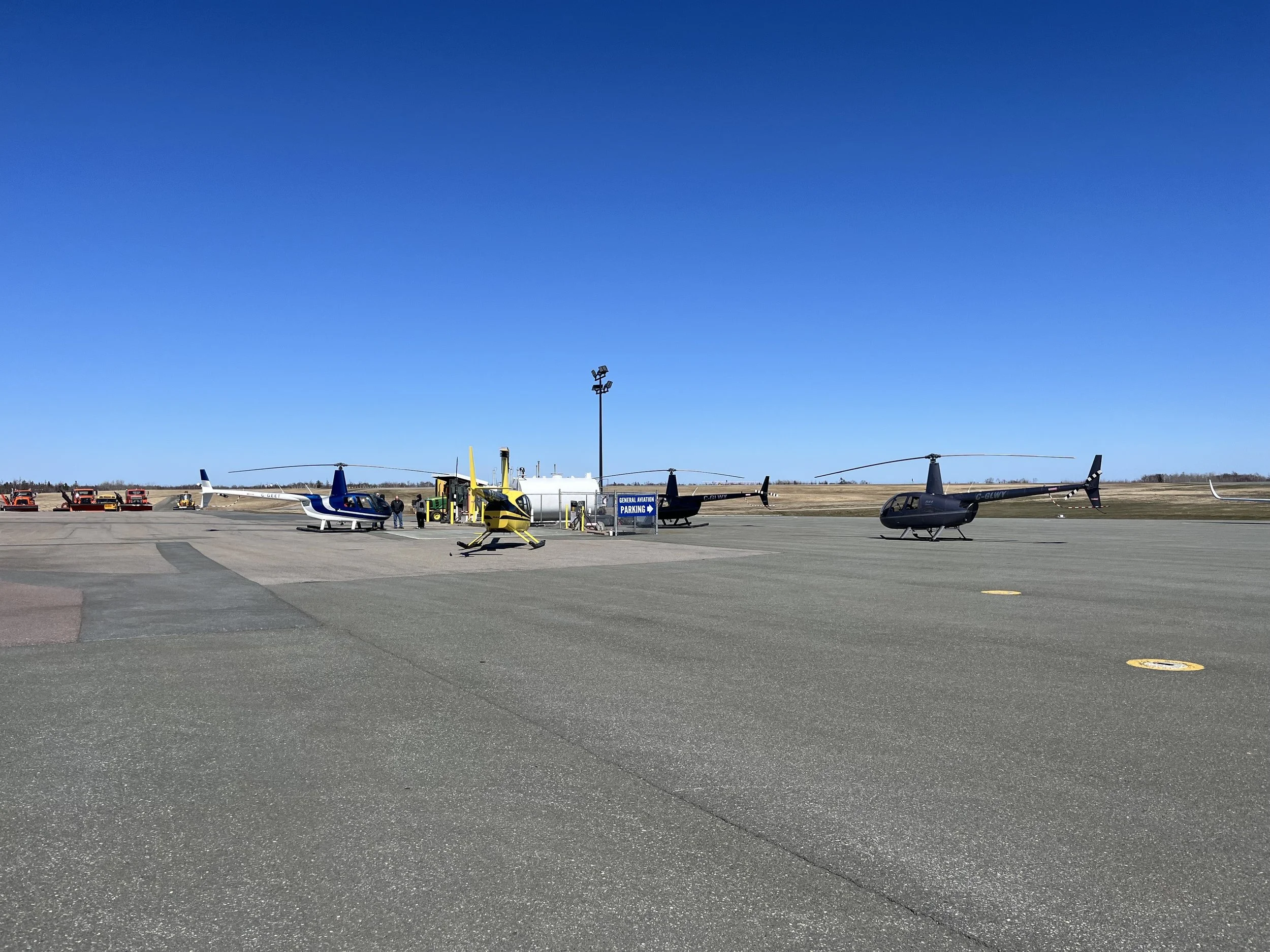 Three helicopters parked on a tarmac at an airport, with a fueling station in the background and a clear blue sky above.
