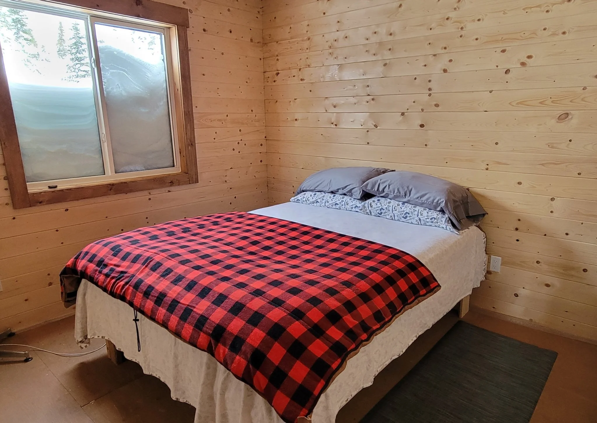 A bedroom with a bed covered in a red and black checkered blanket, two gray pillows, and sheet, in a wooden-paneled room, with a window showing a snowy landscape outside.
