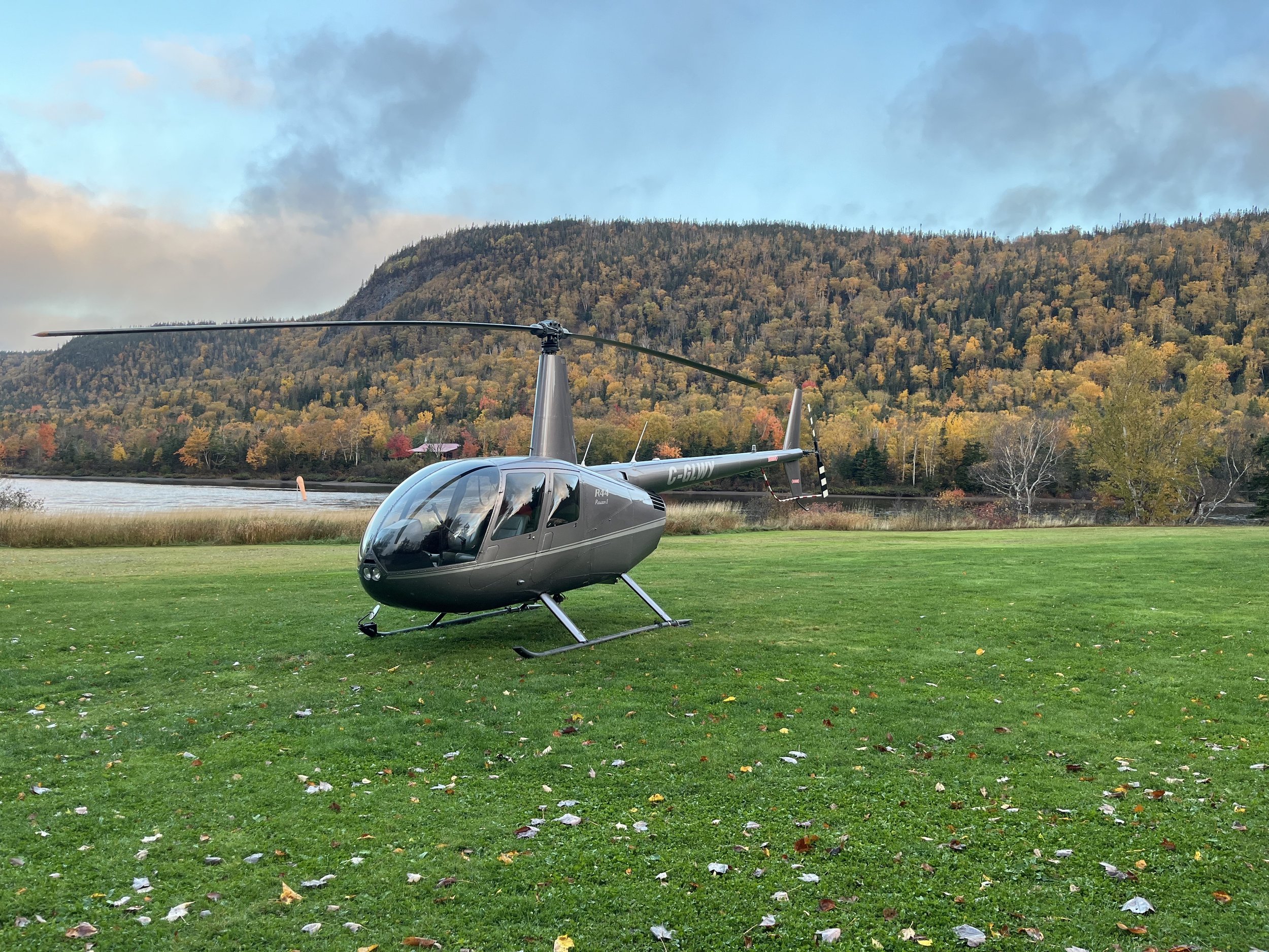 A gray helicopter parked on a grassy field with fall trees, a river, and a mountain in the background.