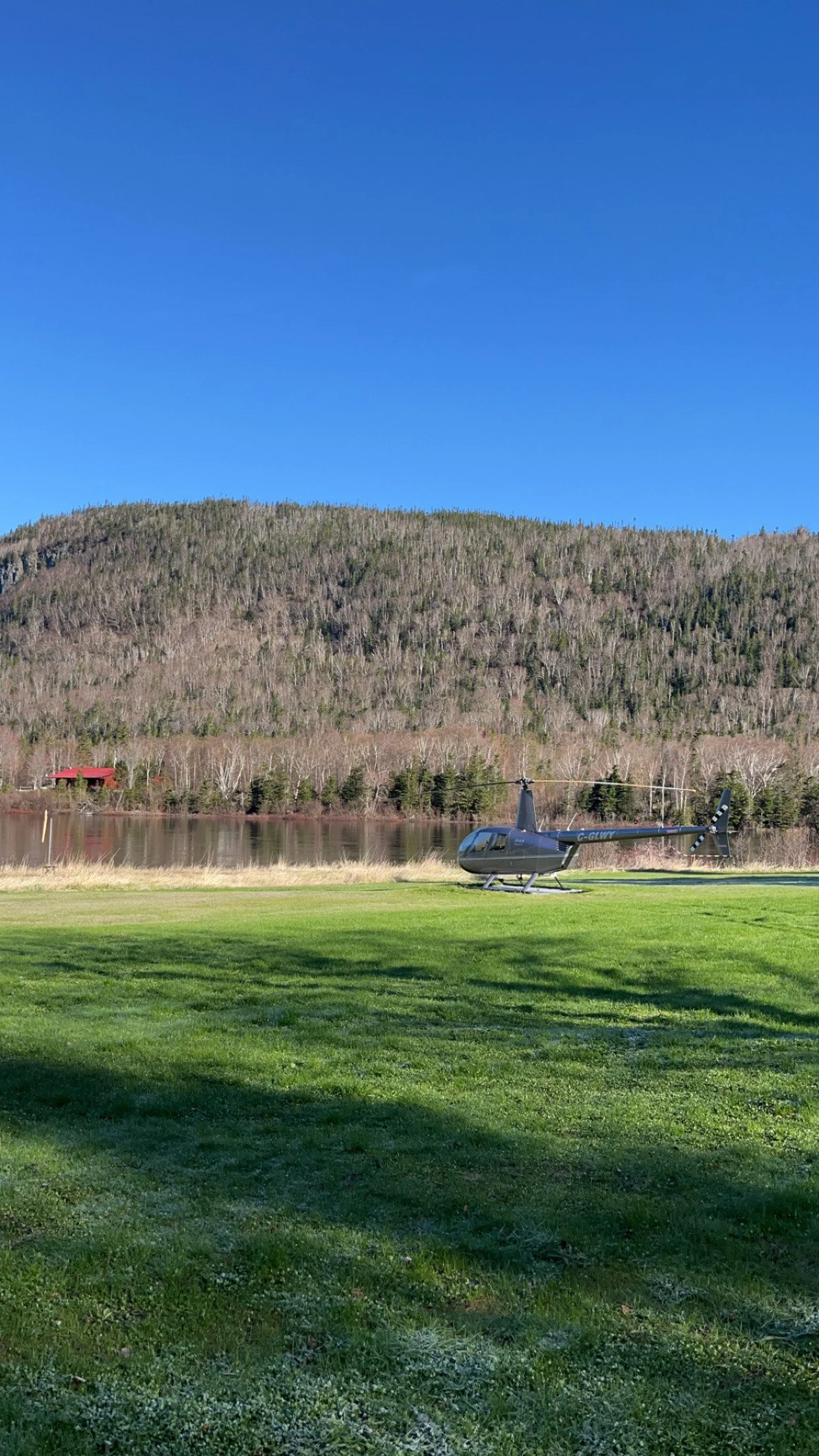 A helicopter parked on a grassy field near a body of water, with a forested hill in the background under a clear blue sky.