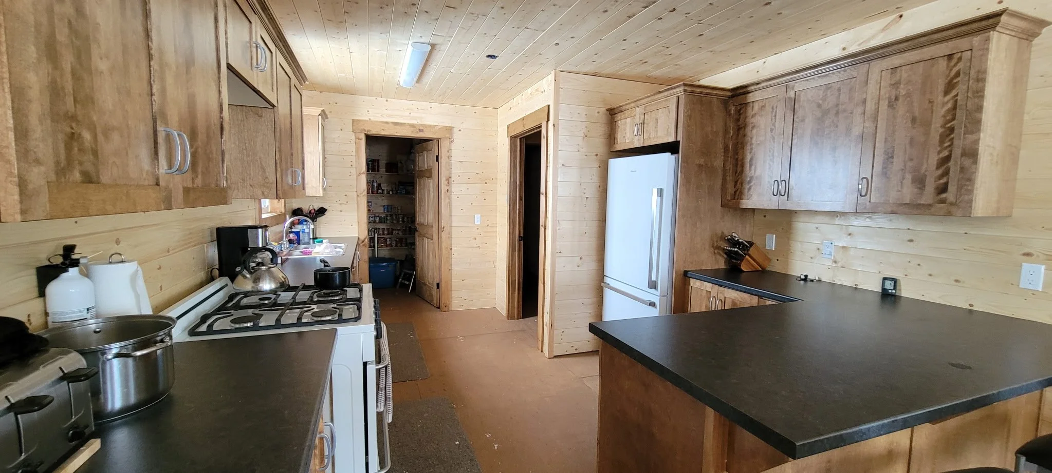A rustic kitchen with wooden cabinets and walls, white appliances including a refrigerator and stove, black countertops, and various kitchen items on the counters. There is a doorway leading to a pantry and another doorway to a different room.