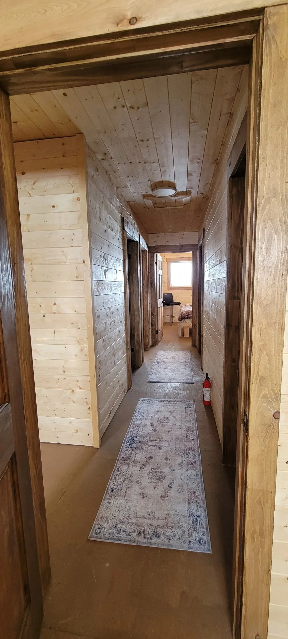A hallway with wooden paneling on the walls and ceiling, with three runners on the floor leading to a room with a window and furniture, and a fire extinguisher on the right side.