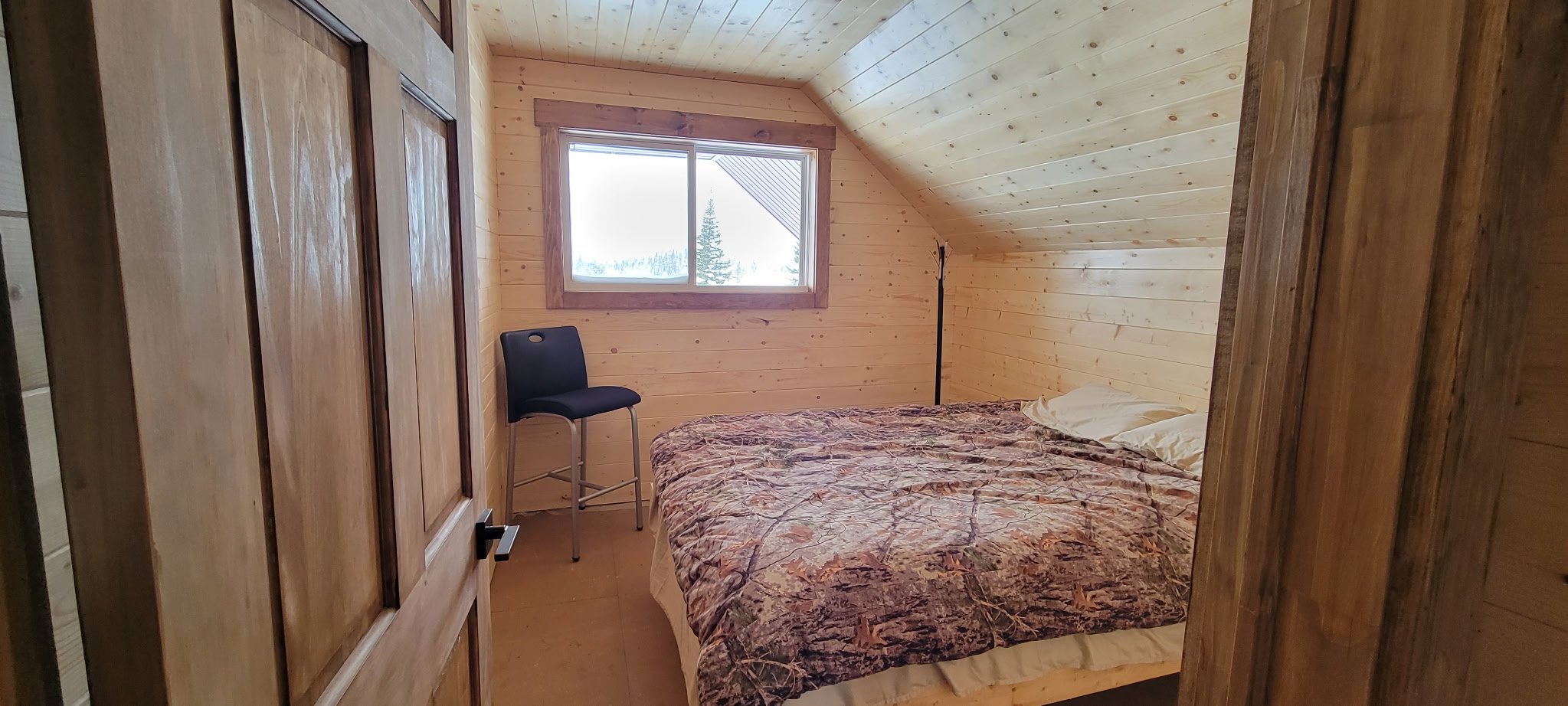 A small wooden bedroom with a slanted ceiling, a window showing an outside view of trees, a black chair, a bed with a camouflage-patterned blanket, and a wooden wardrobe.