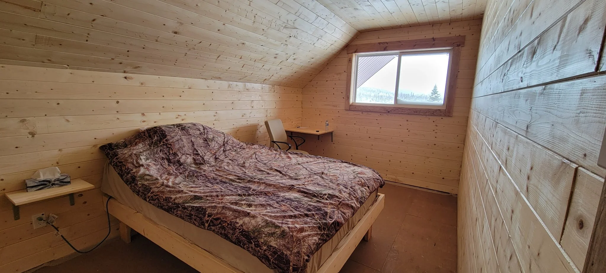 A simple bedroom with wood-paneled walls and sloped ceiling, a bed with camouflage bedding, a small desk with a chair near the window, and a box of tissues on a small shelf.