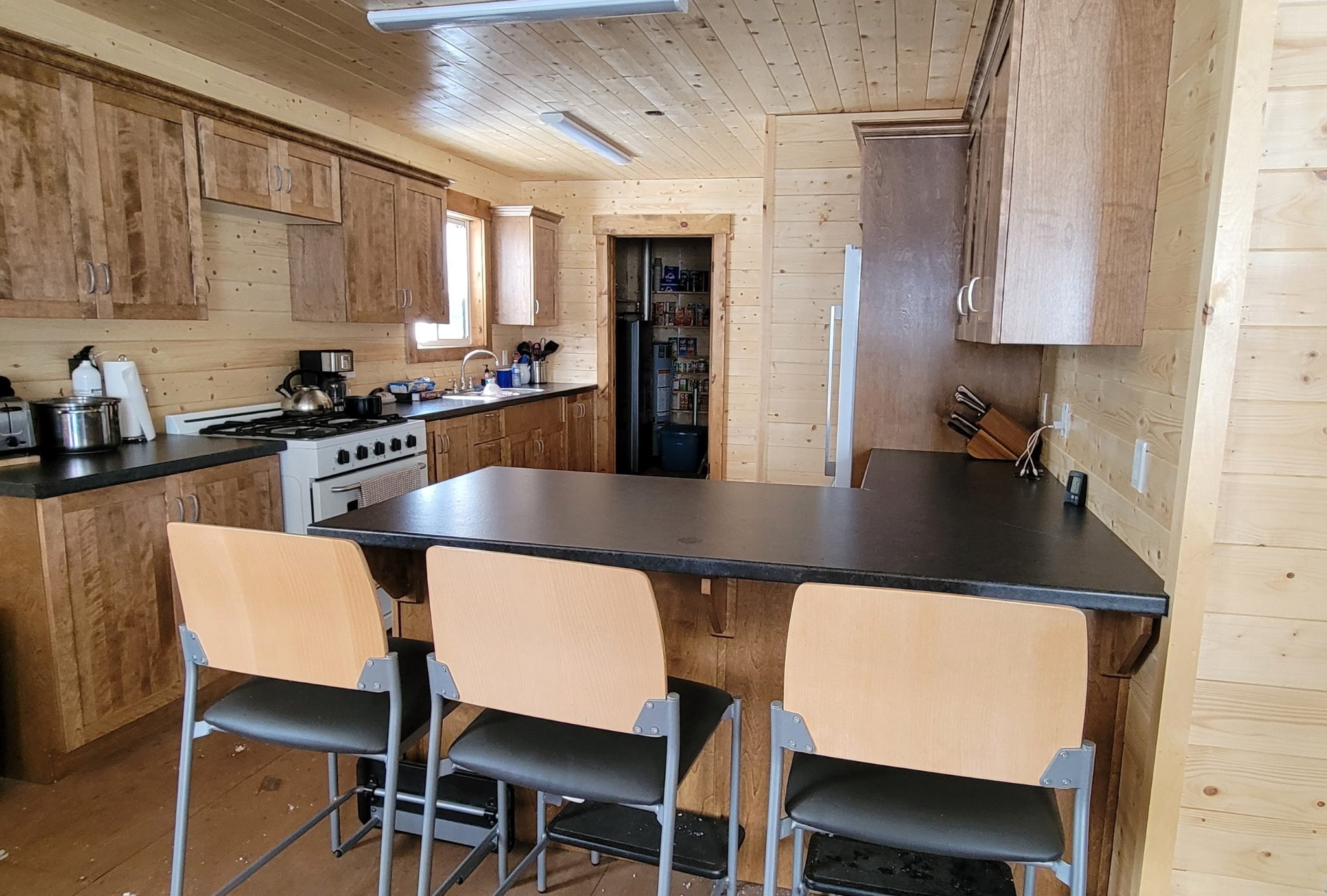 A rustic kitchen with wooden cabinetry, black countertops, and a black kitchen island with three chairs. The kitchen has a stove, sink, and various appliances, with a pantry in the background.