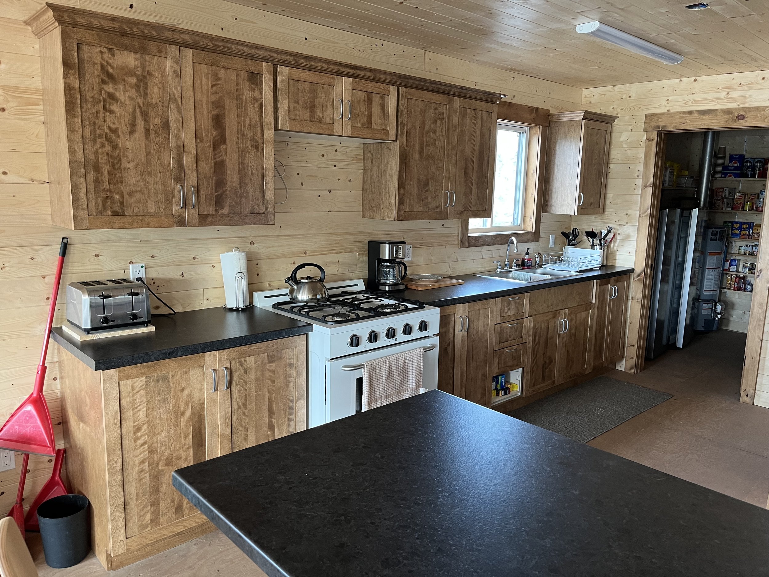 Kitchen with wooden cabinets, black countertops, white stove, coffee maker, toaster, paper towels, kitchen sink, utensils, window, pantry with canned goods and other items.