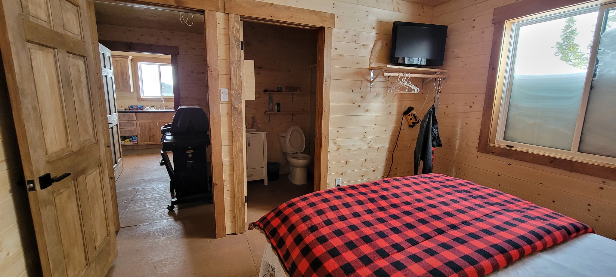 A cozy wooden bedroom with a bed covered in a red and black checkered blanket, a window letting in natural light, a wall-mounted TV, a coat rack with clothes hanging, and an open door leading to a bathroom with a toilet. The room has wooden walls and